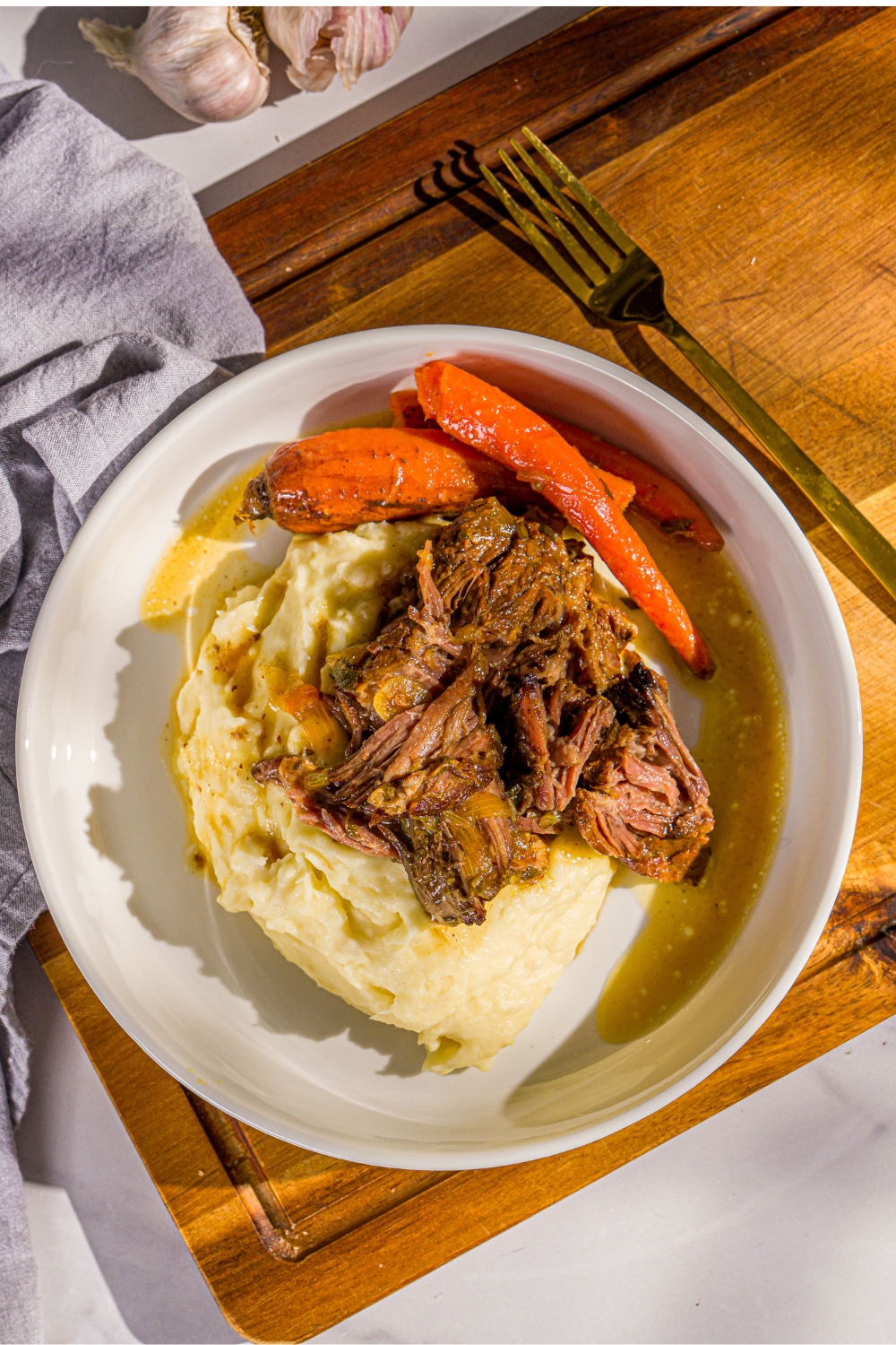 A white plate with apple cider pot roast served over mashed potatoes with carrots. The plate is on a wooden board with a fork and gray cloth napkin.