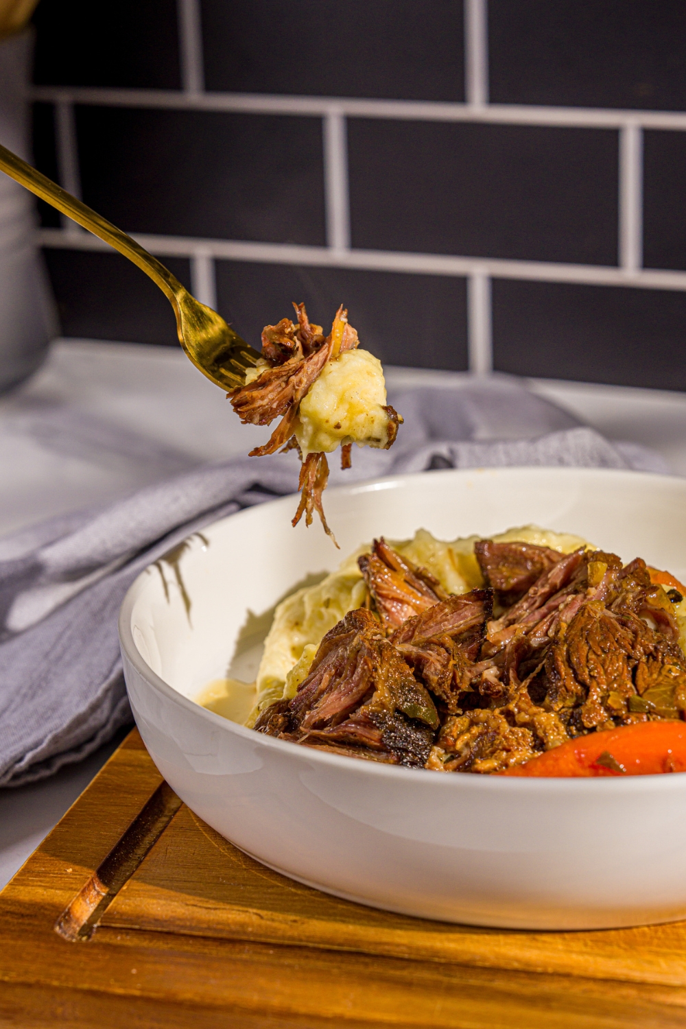 A white plate with apple cider pot roast served over mashed potatoes with carrots. A fork is taking a bite from the plate. The plate is on a wooden board with a fork and gray cloth napkin.
