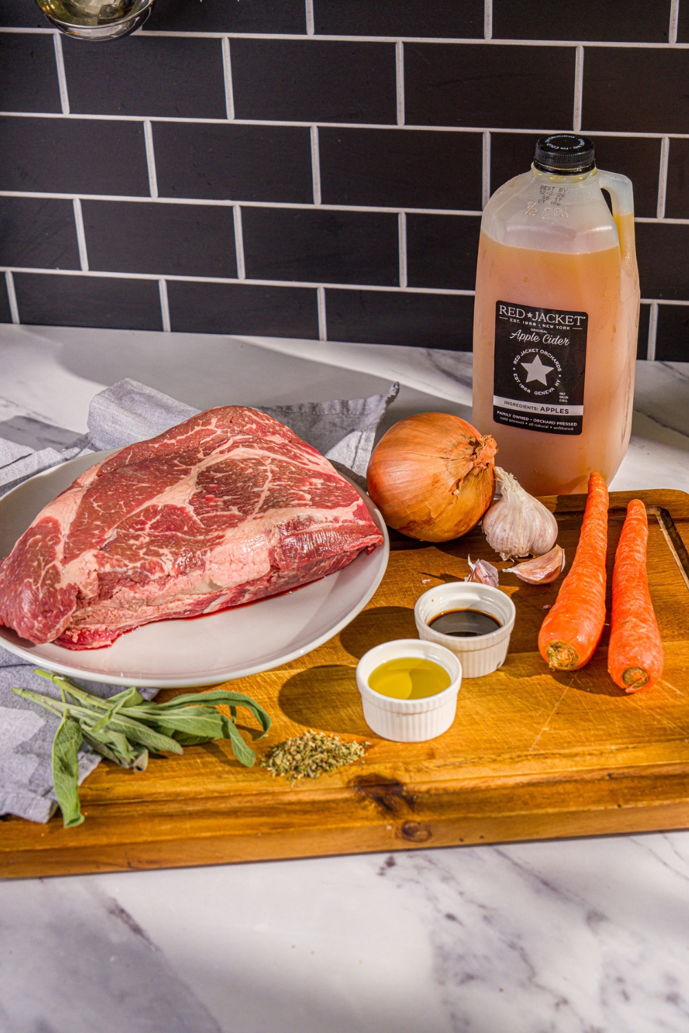A wooden board with ingredients to make apple cider pot roast including a chuck roast, apple cider, onion, carrots, oil, sage, Worcestersire sauce, and garlic.