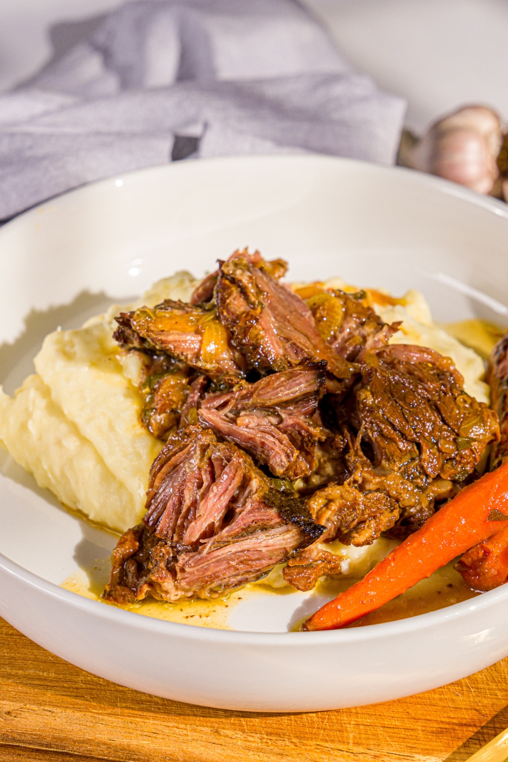 A white plate with apple cider pot roast served over mashed potatoes with carrots. The plate is on a wooden board with a gray cloth napkin.