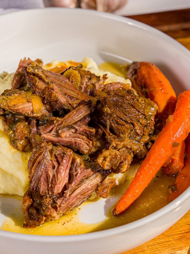 A white plate with apple cider pot roast served over mashed potatoes with carrots. The plate is on a wooden board with a gray cloth napkin.