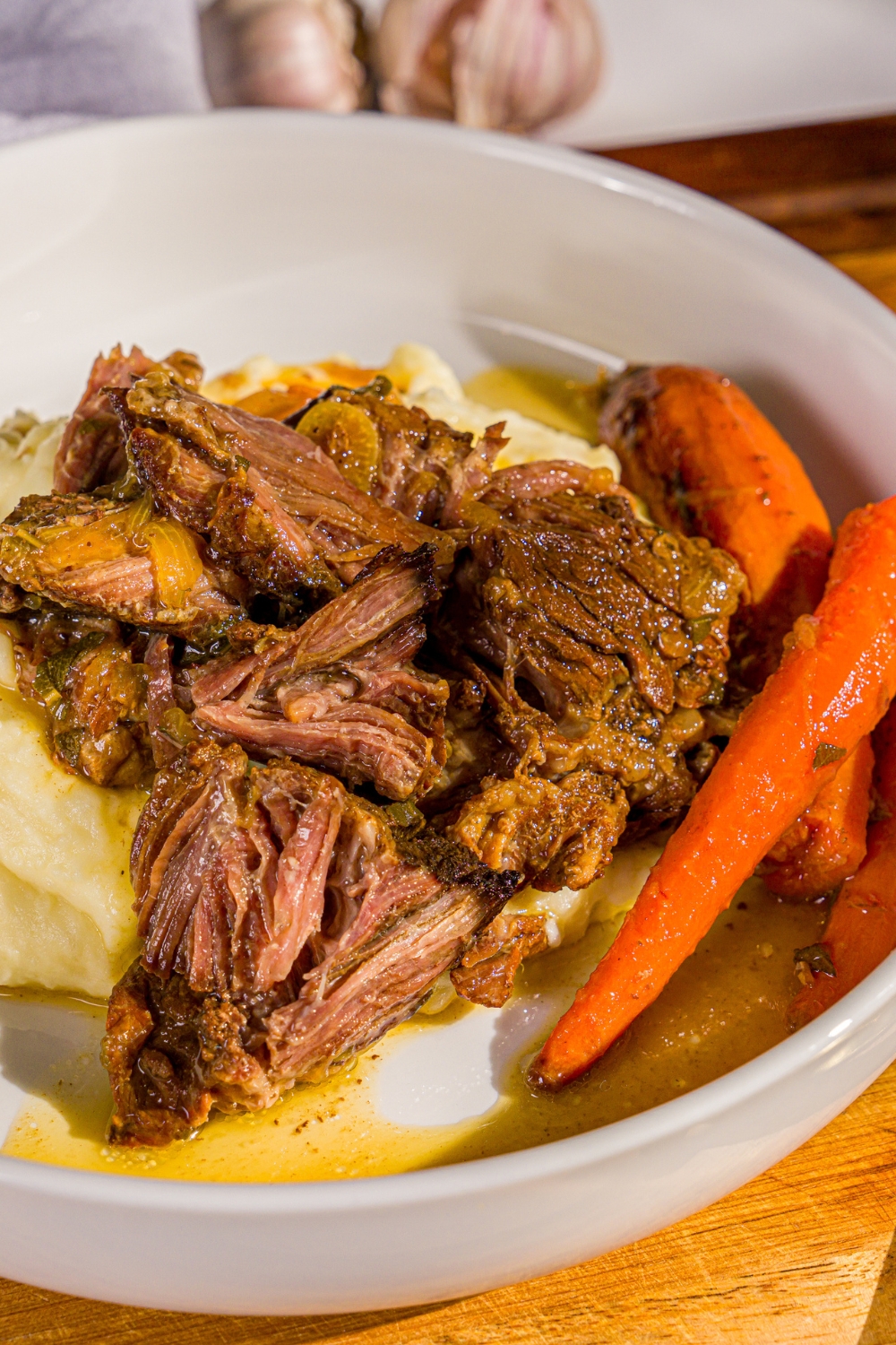 A white plate with apple cider pot roast served over mashed potatoes with carrots. The plate is on a wooden board with a gray cloth napkin.