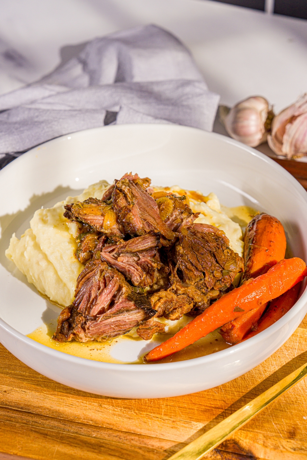 A white plate with apple cider pot roast served over mashed potatoes with carrots. The plate is on a wooden board with a gray cloth napkin.
