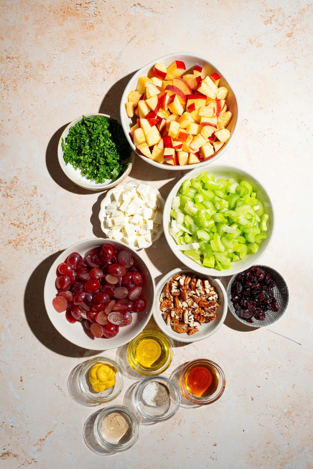 A tan counter with several bowls containing ingredients to make apple crunch salad including apples, pecans, grapes, parsley, feta cheese, dried cranberries, celery, olive oil, apple cider vinegar, dijon mustard, and more.