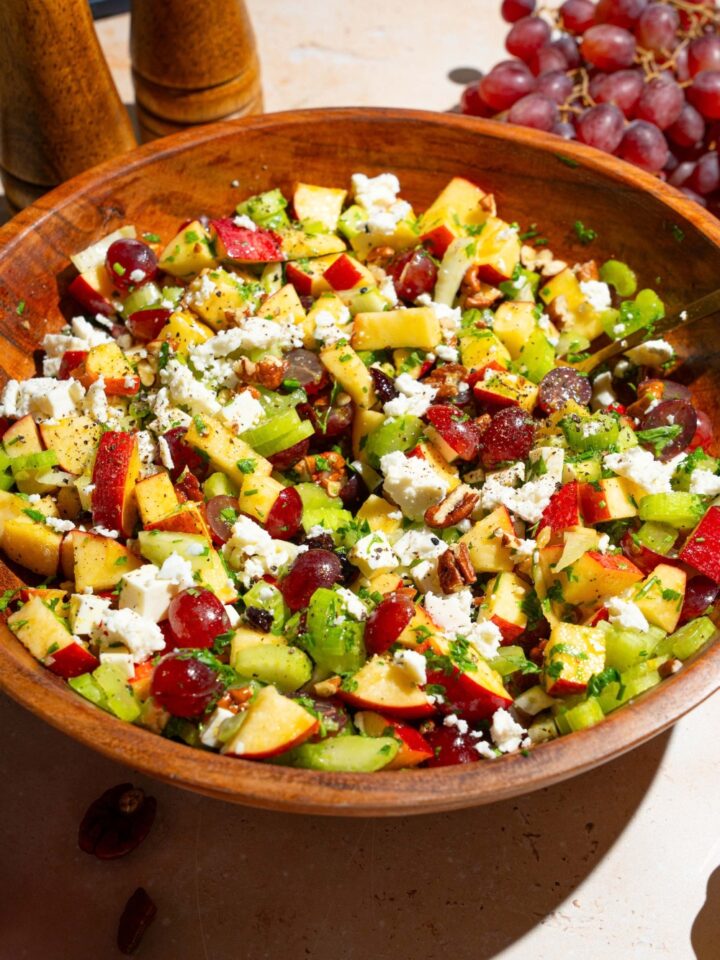 A wooden bowl with apple crunch salad including apples, celery, pecans, feta cheese, parsley, and grapes tossed in a maple dijon dressing. The bowl is on a tan counter with grapes.