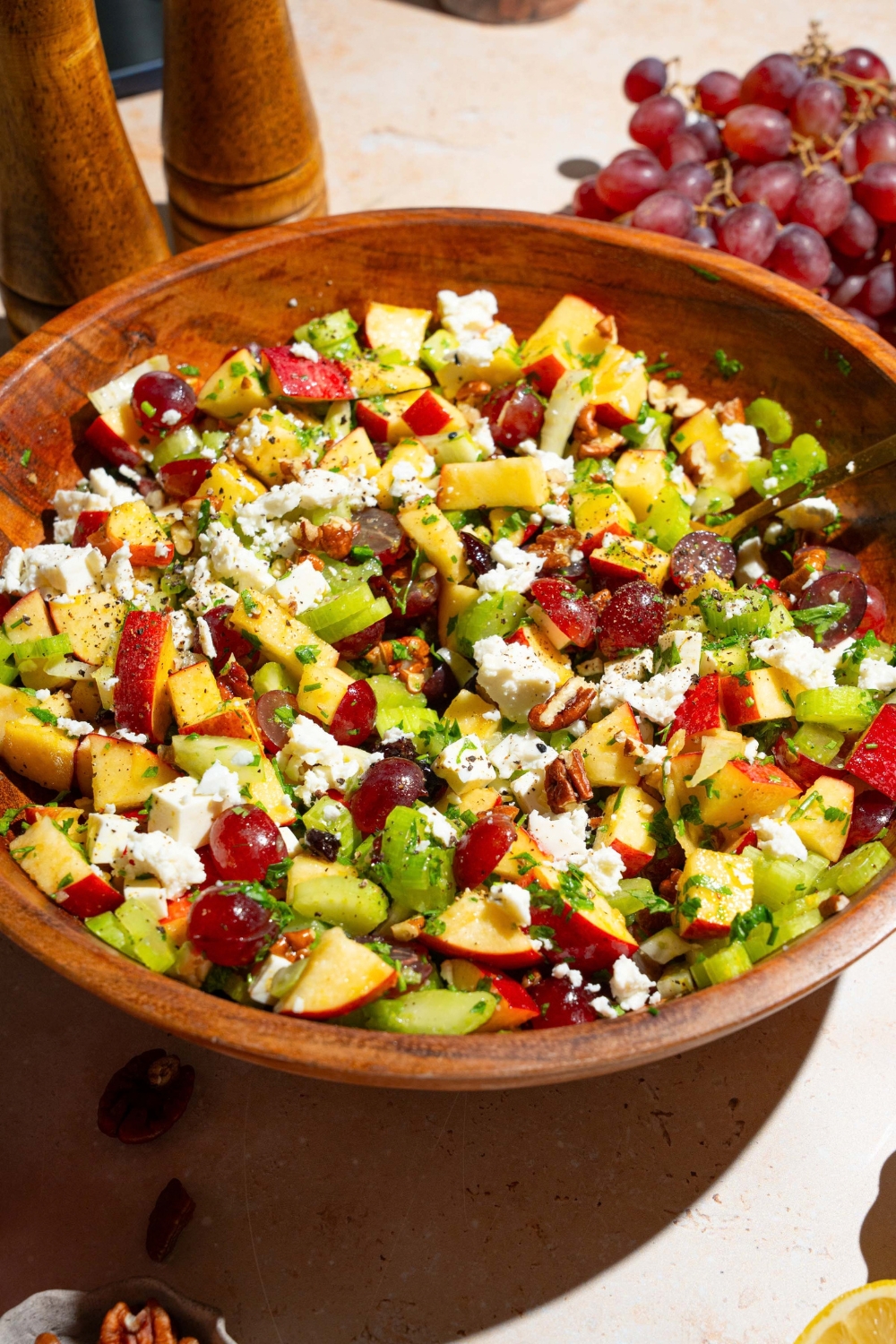 A wooden bowl with apple crunch salad including apples, celery, pecans, feta cheese, parsley, and grapes tossed in a maple dijon dressing. The bowl is on a tan counter with grapes.