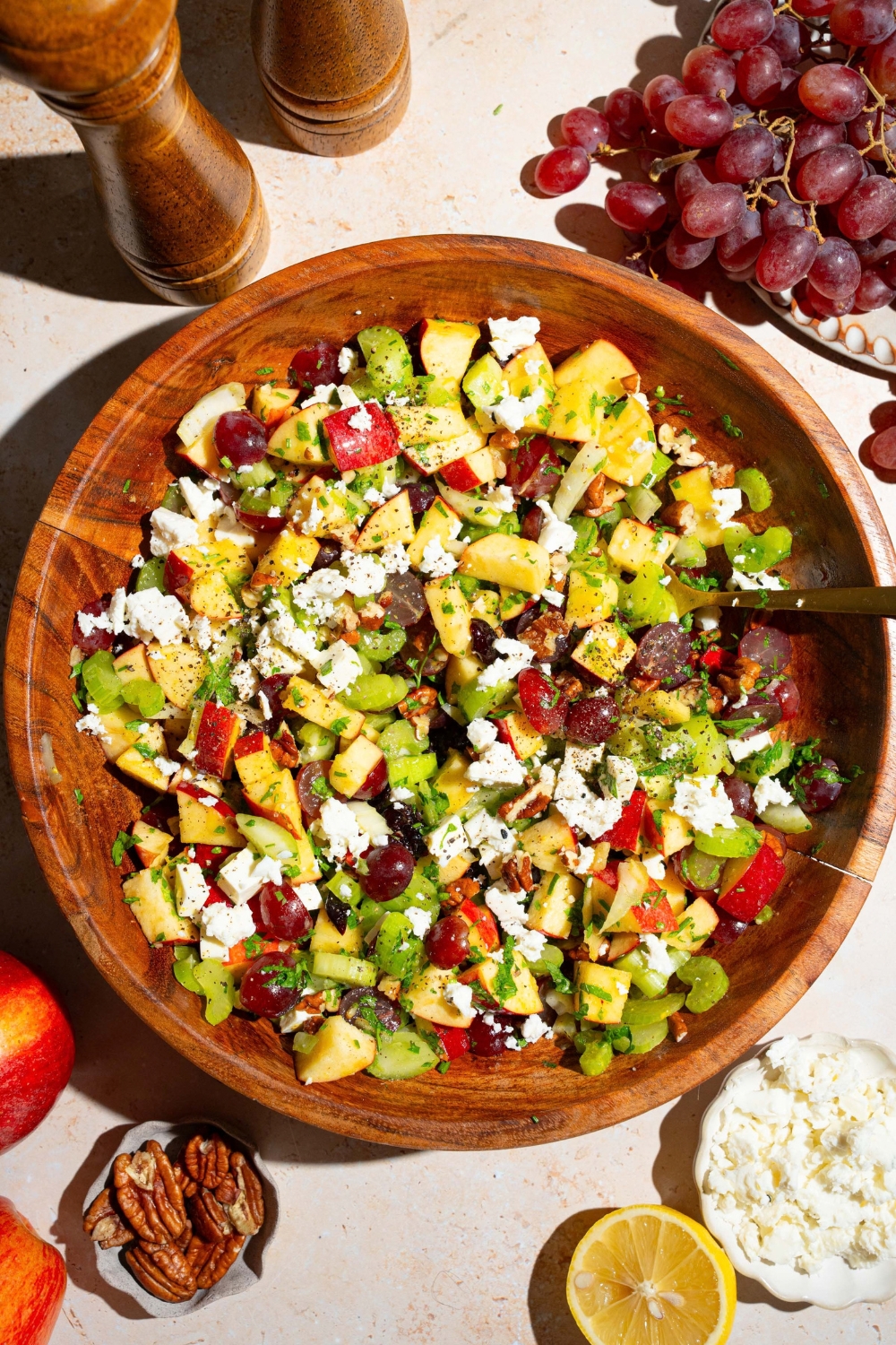 A wooden bowl with apple crunch salad including apples, celery, pecans, feta cheese, parsley, and grapes tossed in a maple dijon dressing. There is a fork in the bowl. The bowl is on a tan counter with grapes, pecans, and lemon.