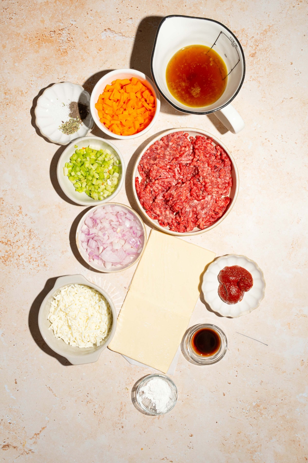 A tan counter with several bowls containing ingredients to make beef pot pie including pie crust, ground beef, carrots, celery, onion, tomato paste, beef broth, cornstarch, shredded mozzarella, and more.
