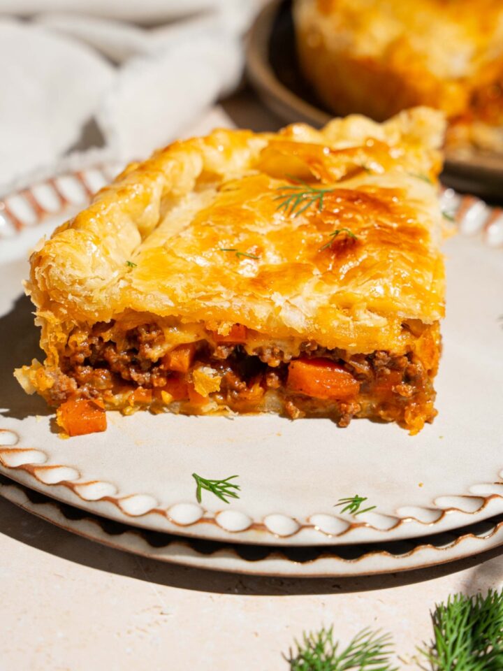 Two plates stacked together with a slice of beef pot pie on top. The pie is garnished with fresh herbs. The plates are a tan counter with a whole beef pot pie.
