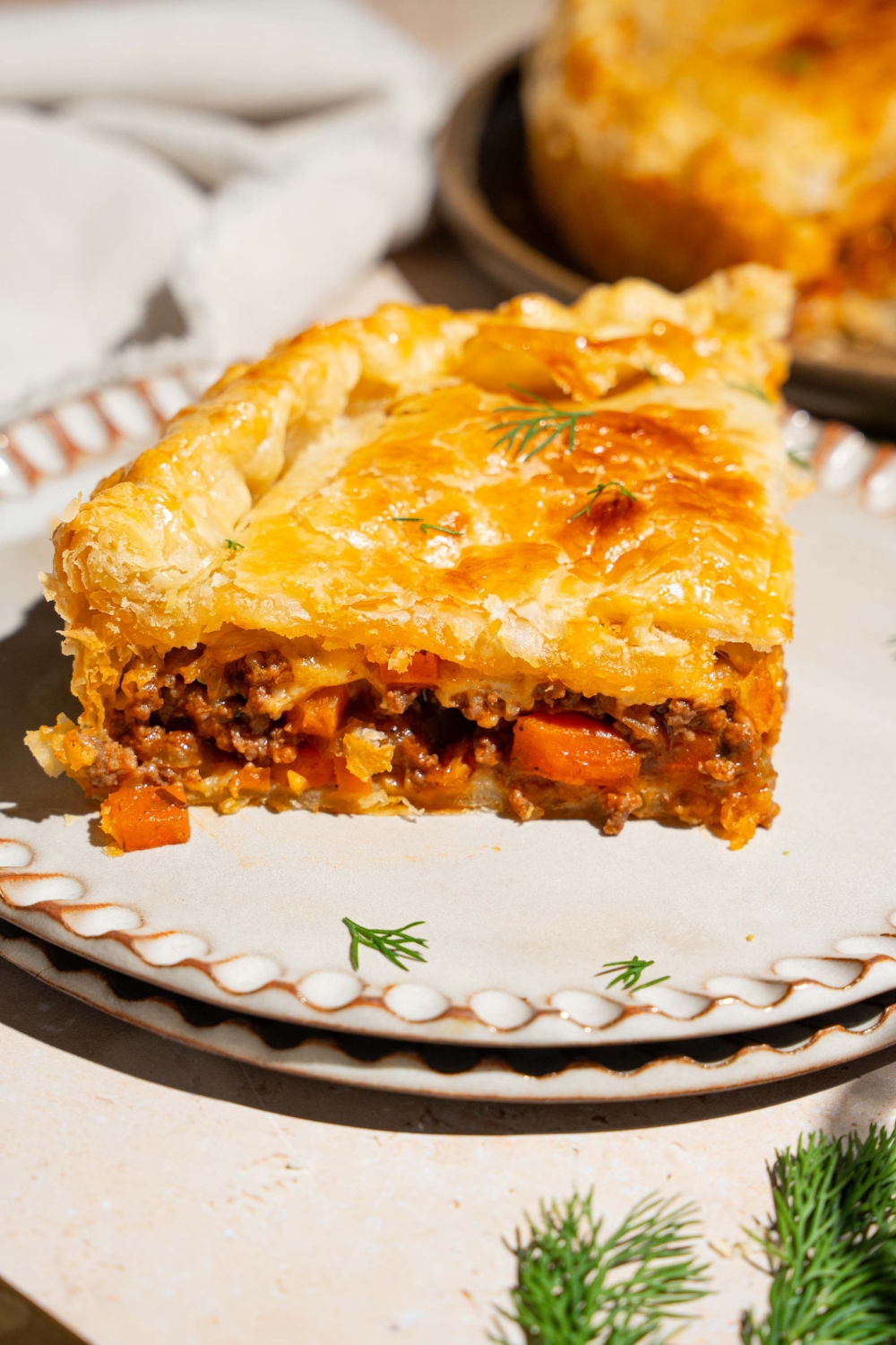 Two plates stacked together with a slice of beef pot pie on top. The pie is garnished with fresh herbs. The plates are a tan counter with a whole beef pot pie.