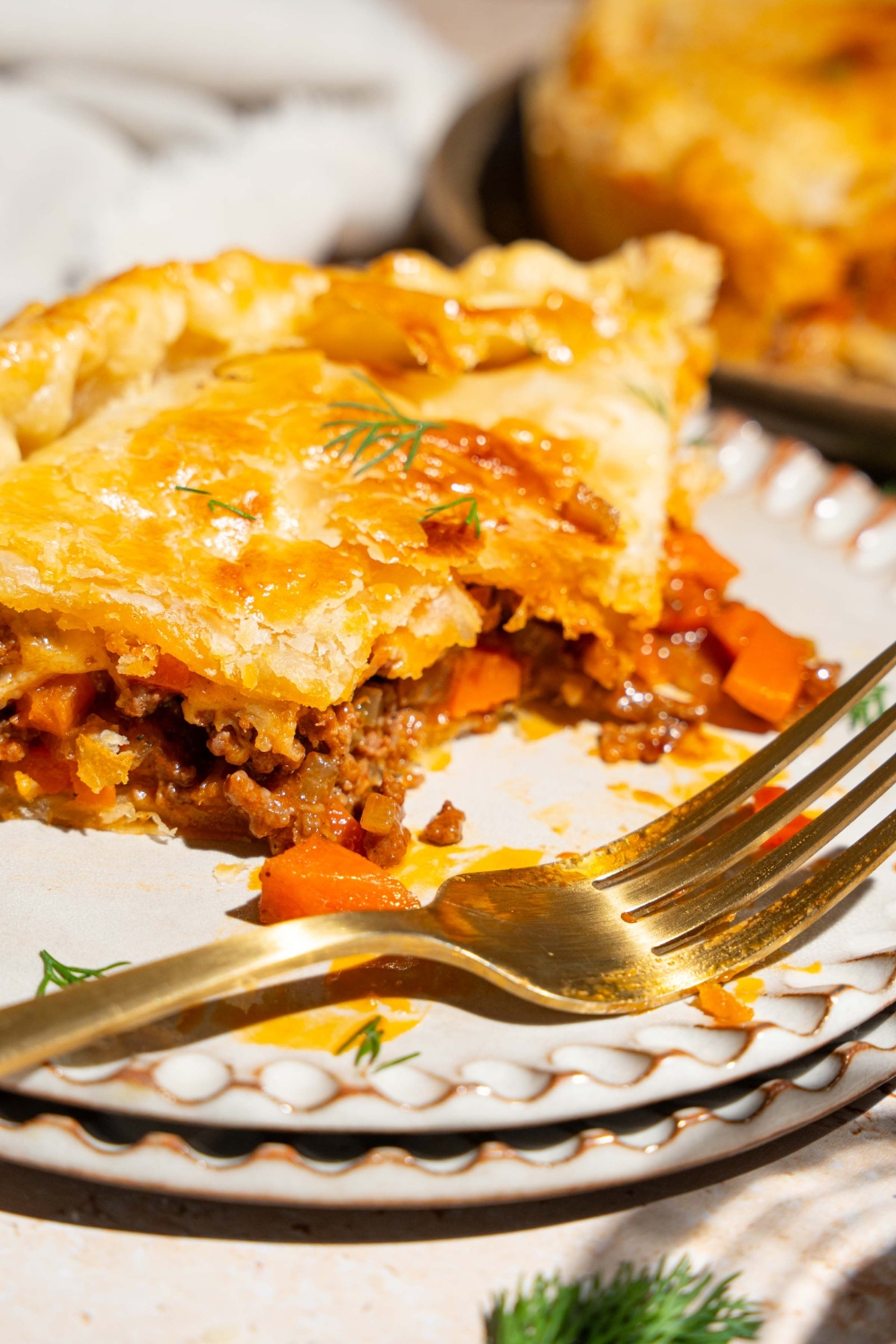 Two plates stacked together with a slice of beef pot pie on top. The pie is garnished with fresh herbs. A bite is taken from slice. There is a fork on the plate. The plates are a tan counter with a whole beef pot pie.