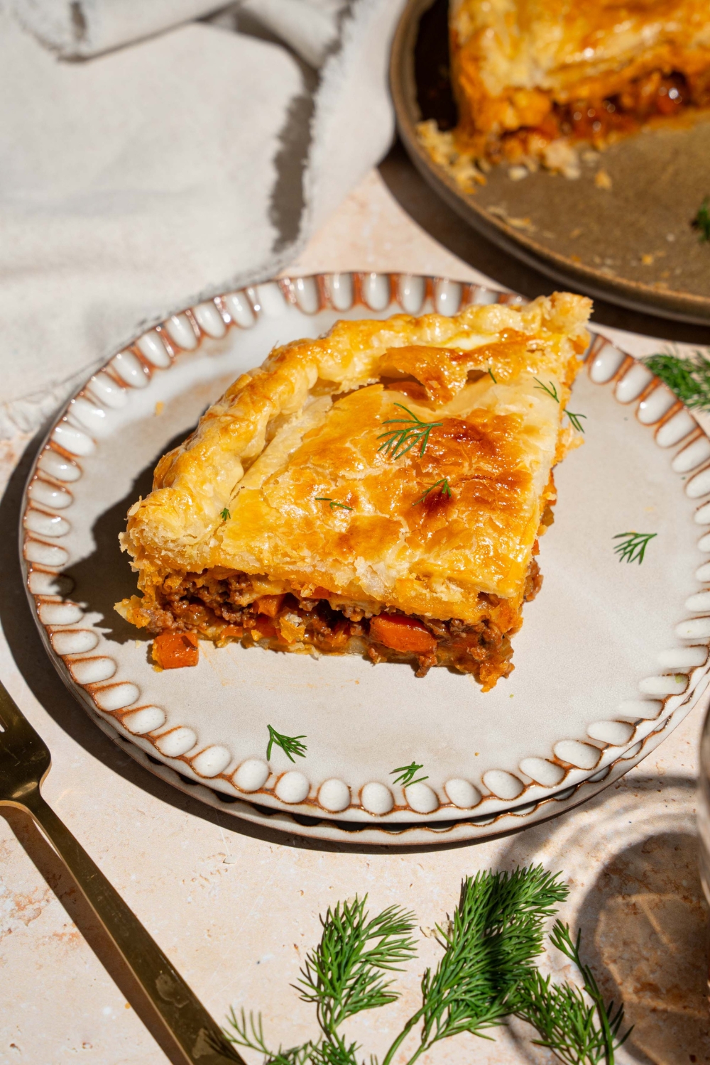 Two plates stacked together with a slice of beef pot pie on top. The pie is garnished with fresh herbs. The plates are a tan counter with a whole beef pot pie.