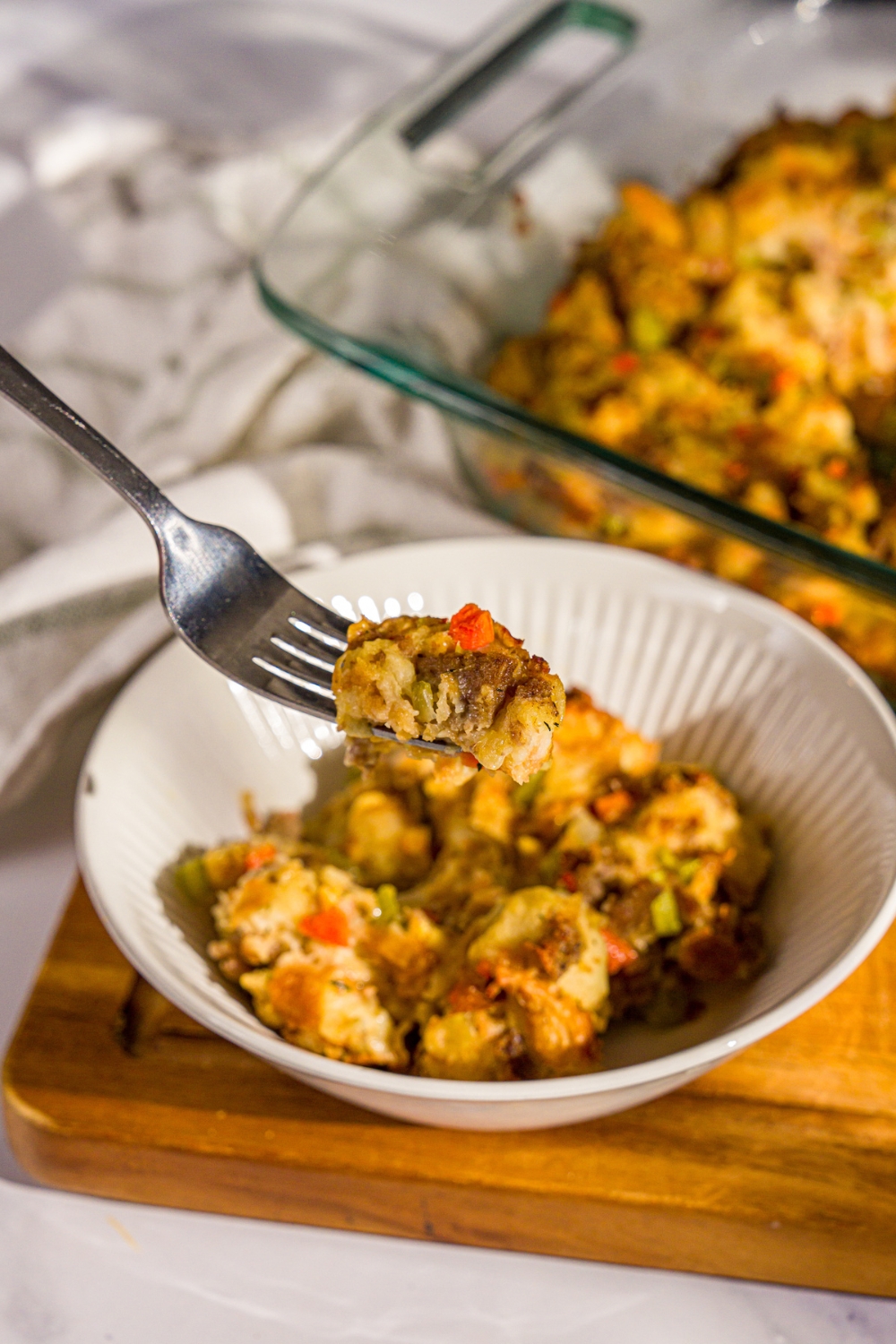 A bowl of White Castle stuffing with a fork taking a bite from the bowl. The bowl is on a wooden board with a baking dish of stuffing.