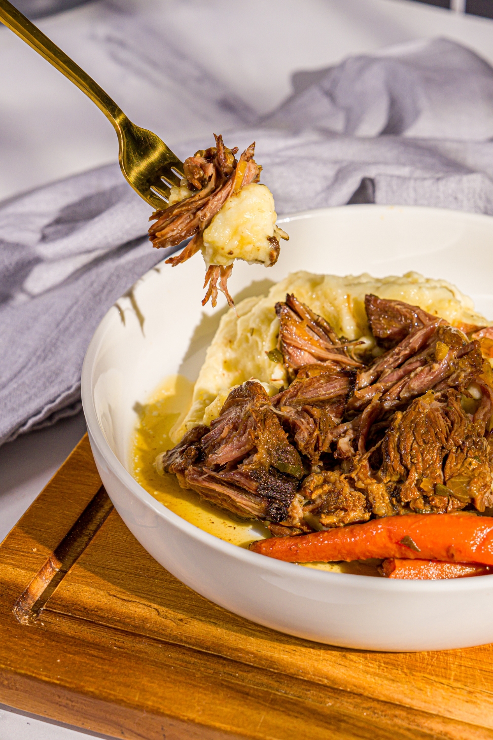 A white plate with apple cider pot roast served over mashed potatoes with carrots. A fork is taking a bite from the plate. The plate is on a wooden board with a gray cloth napkin.