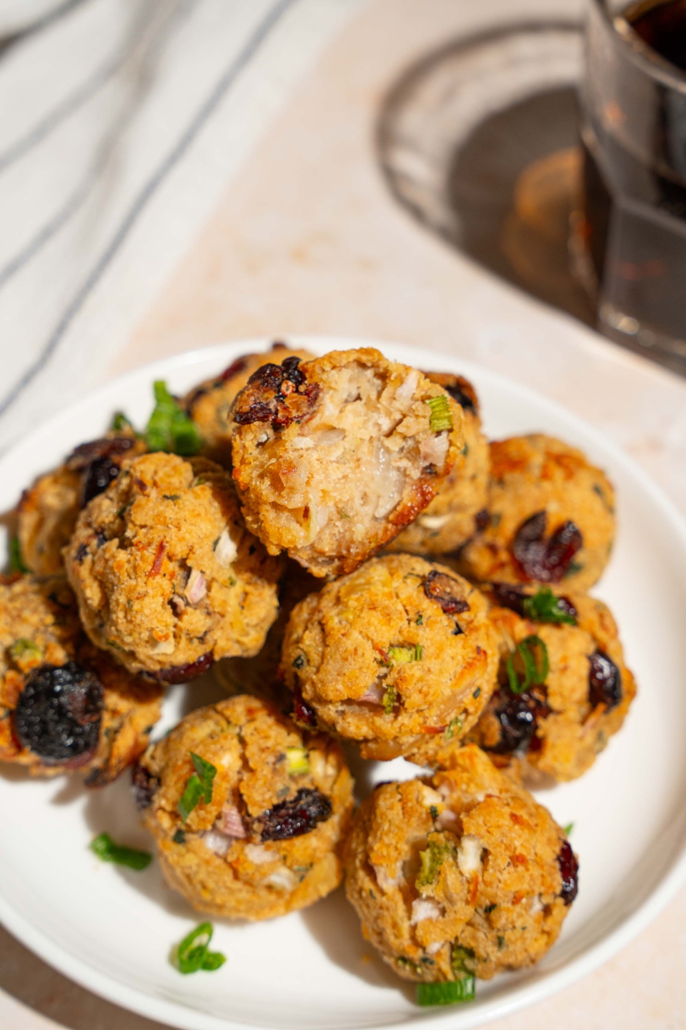 A white plate with cranberry turkey stuffing balls garnished with sliced green onions. One of the balls has a bite removed. The plate is on a tan counter with a white striped napkin.