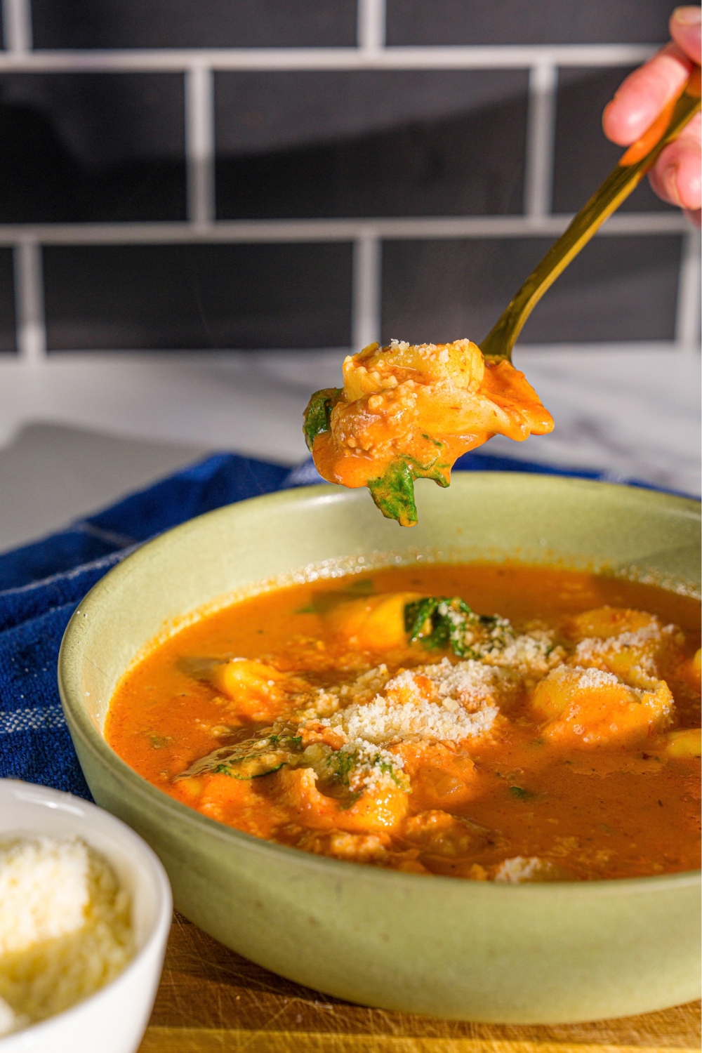 A bowl of creamy tomato tortellini soup topped with shredded parmesan. A spoon is taking a bite from the bowl. The bowl is on a wooden board with a small bowl of cheese and a blue cloth napkin.