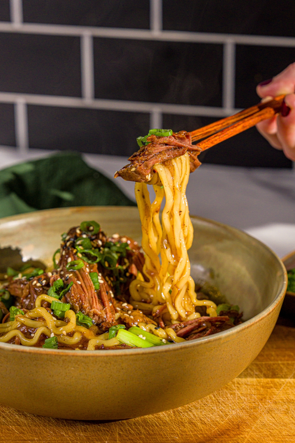 A bowl of crockpot Mongolian beef served with noodles and garnished with sliced green onions and sesame seeds. A pair of chopsticks are pulling a bite from the bowl. The bowl is on a wooden board.