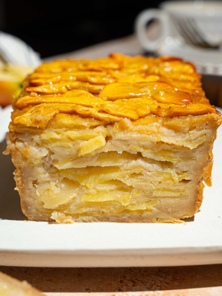A white platter with a whole invisible apple cake. A slice of the cake is removed, showing layers of apple cake.