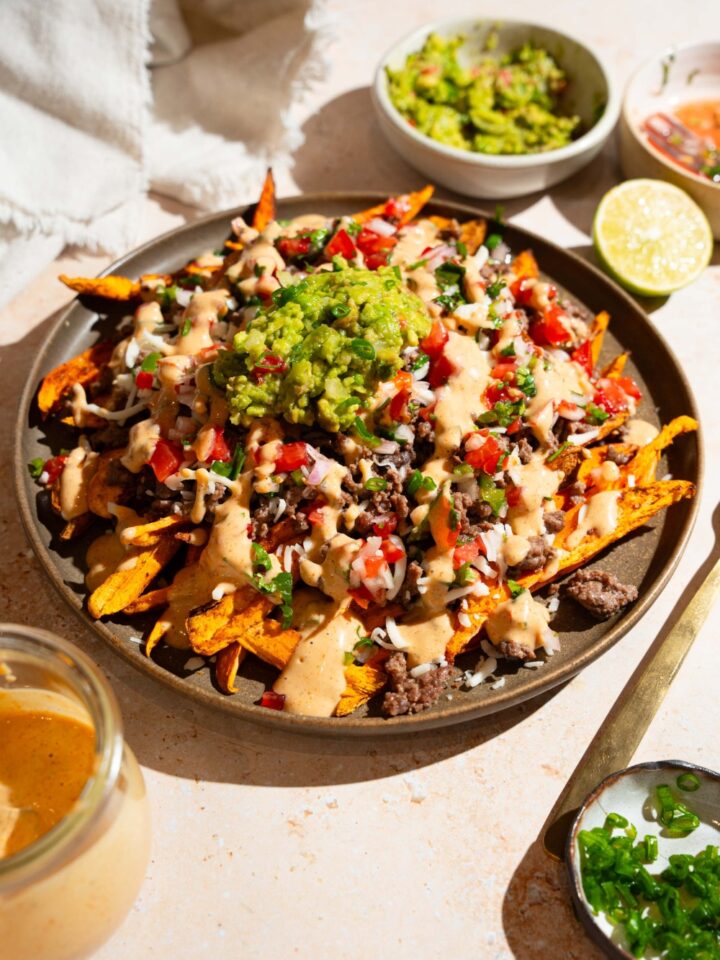 A plate of loaded sweet potato fries topped with ground beef, chipotle ranch, pico de gallo, shredded cheese, and guacamole. The plate is on a tan counter with small bowls of toppings.