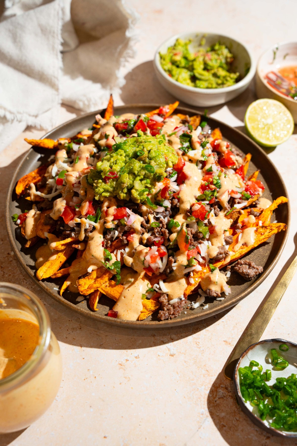 A plate of loaded sweet potato fries topped with ground beef, chipotle ranch, pico de gallo, shredded cheese, and guacamole. The plate is on a tan counter with small bowls of toppings.