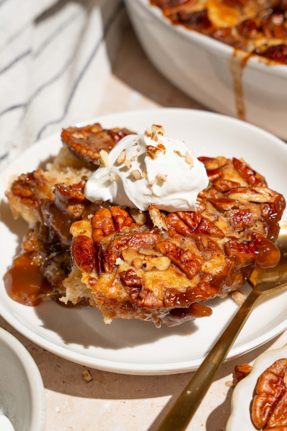 A white plate with a slice of pecan pie cobbler topped with a dollop of whipped cream with chopped pecans. There is a spoon on the plate. The plate is on a tan counter with a casserole dish of cobbler and a white striped napkin.