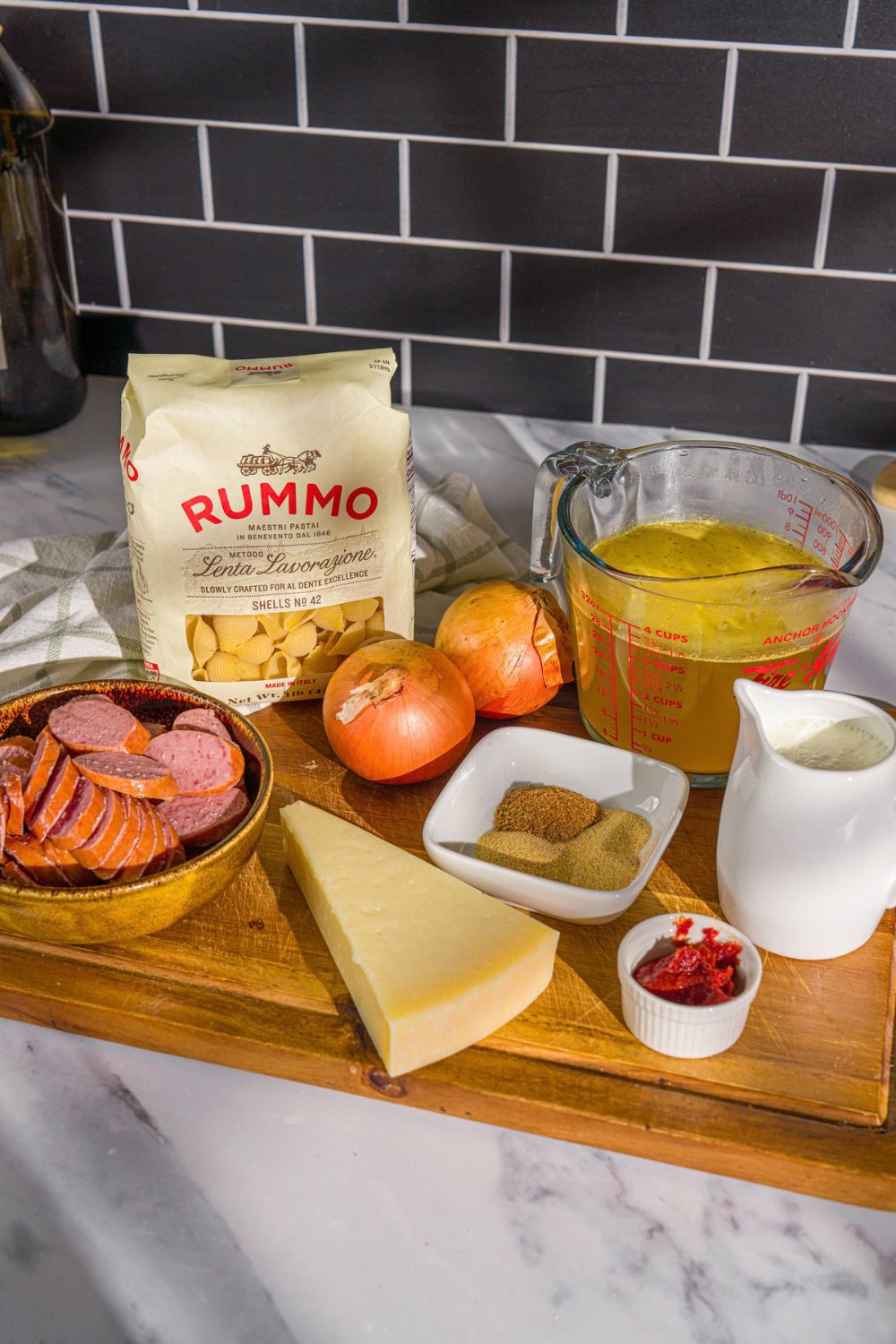 A wooden board with ingredients to make cajun sausage pasta including sausage, onion, pasta, chicken broth, cream, parmesan cheese, tomato paste, and seasonings.