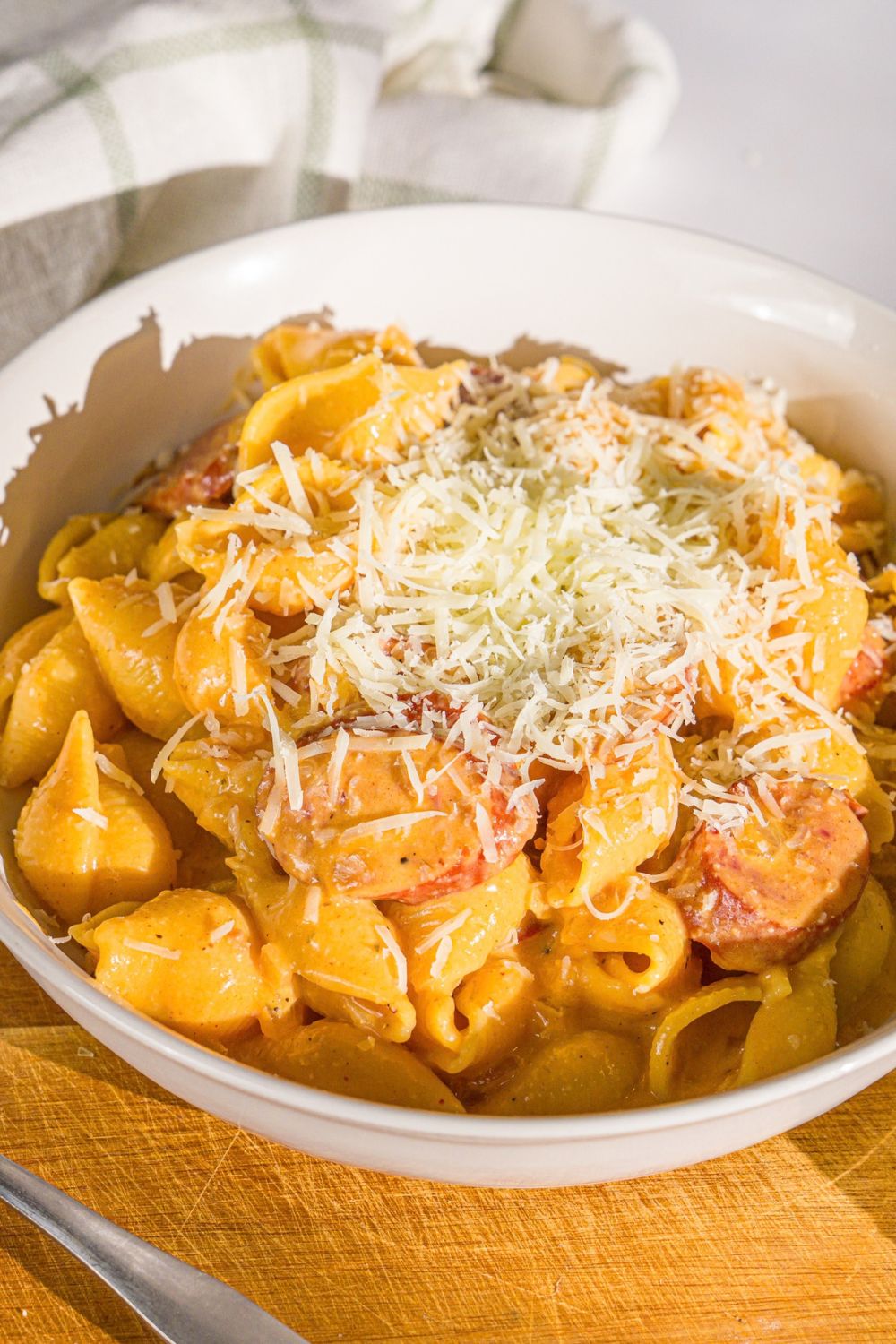 A white bowl with cajun sausage pasta topped with shredded parmesan cheese. The bowl is on a wooden board with a white cloth napkin.