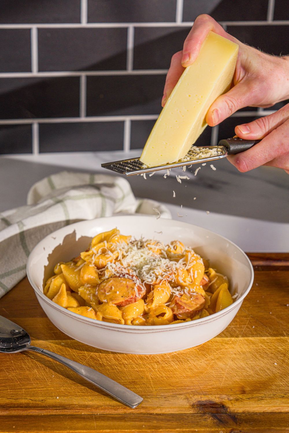 A white bowl with cajun sausage pasta. Parmesan cheese is being grated over the pasta. The bowl is on a wooden board.