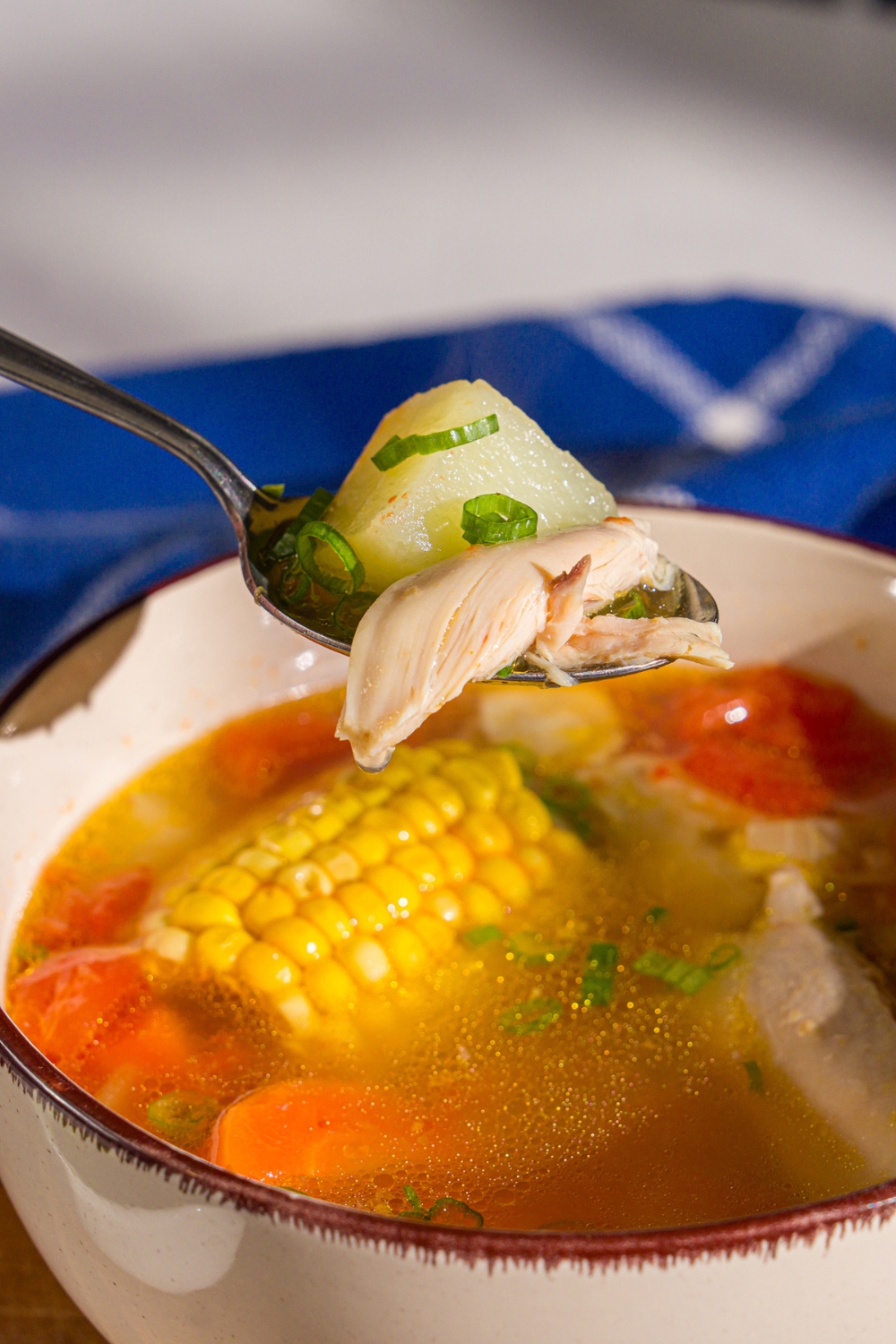 A spoon with a bite of ABC soup with a bowl of ABC soup with chicken, carrots, and corn on the cob garnished with sliced green onion.