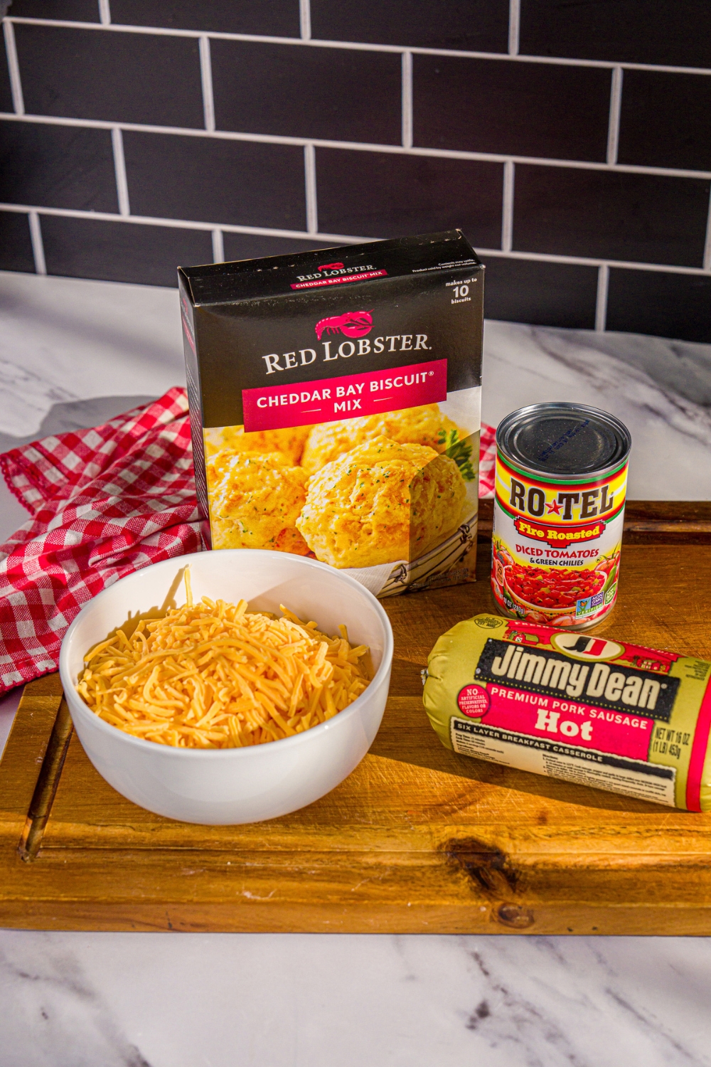 A wooden board with ingredients to make cheddar bay biscuit sausage balls including red lobster cheddar bay biscuit mix, shredded cheese, rotel, and jimmy dean sausage.
