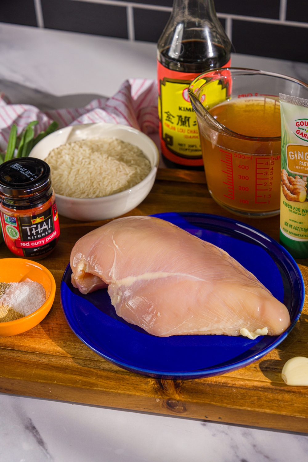 A wooden board with ingredients to make chicken broth bowls including chicken breast, rice, red curry paste, garlic, chicken broth, garlic paste, soy sauce, and more.