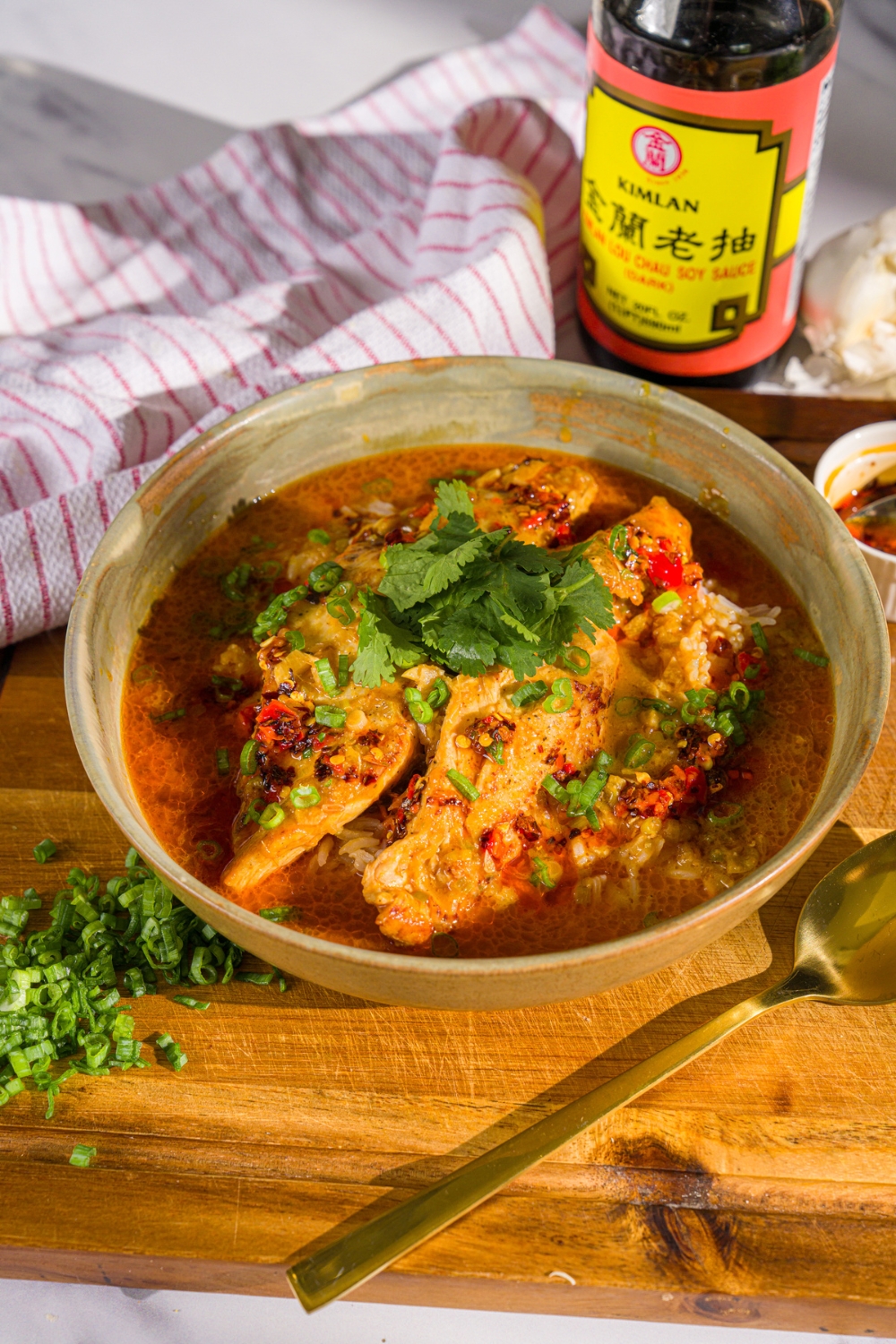 A bowl of thai coconut chicken bowl garnished with sliced green onion, cilantro, and chili oil. The bowl is on a wooden board with a spoon, chopped green onion, and a white cloth napkin.
