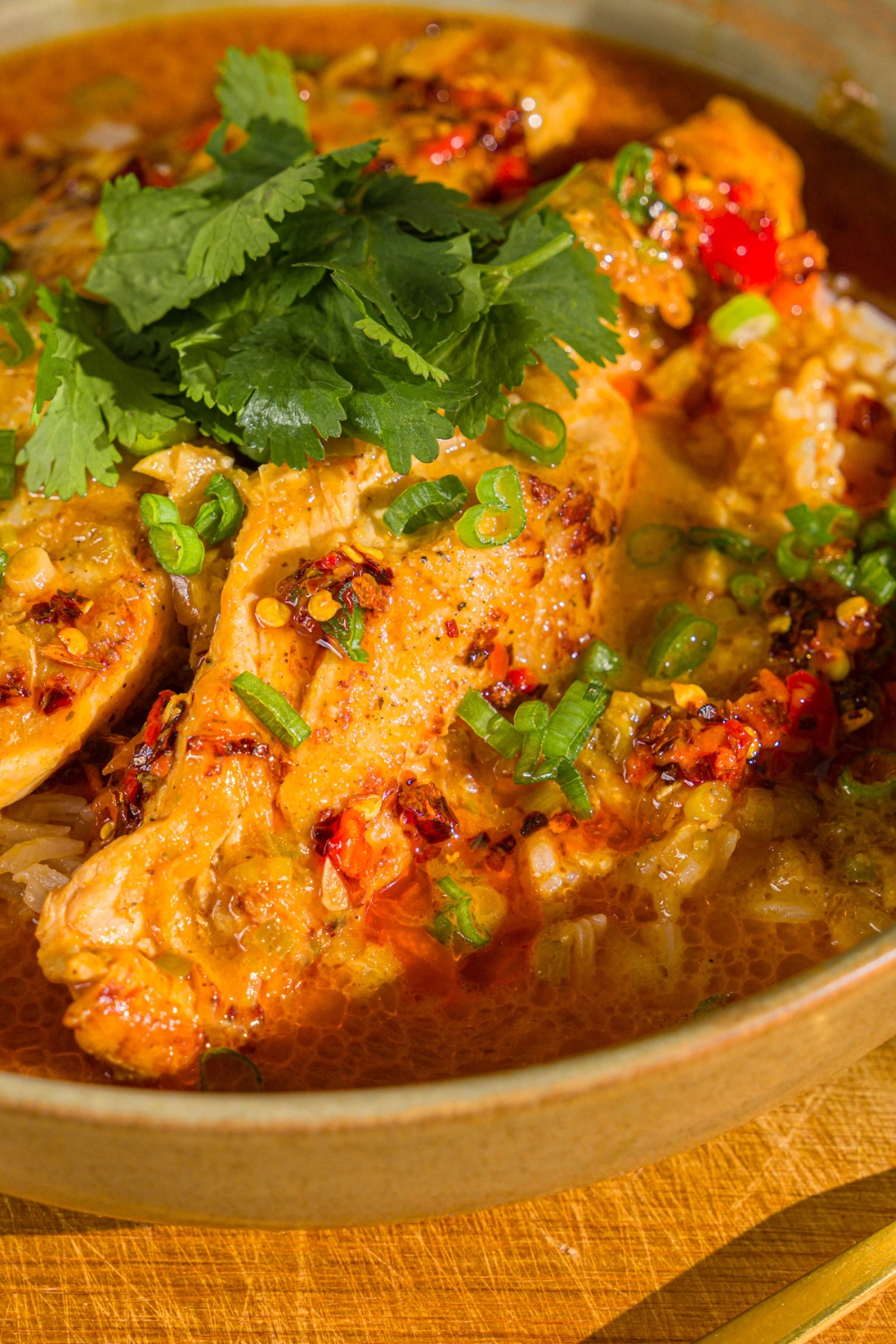 A bowl of thai coconut chicken bowl garnished with sliced green onion, cilantro, and chili oil. The bowl is on a wooden board.