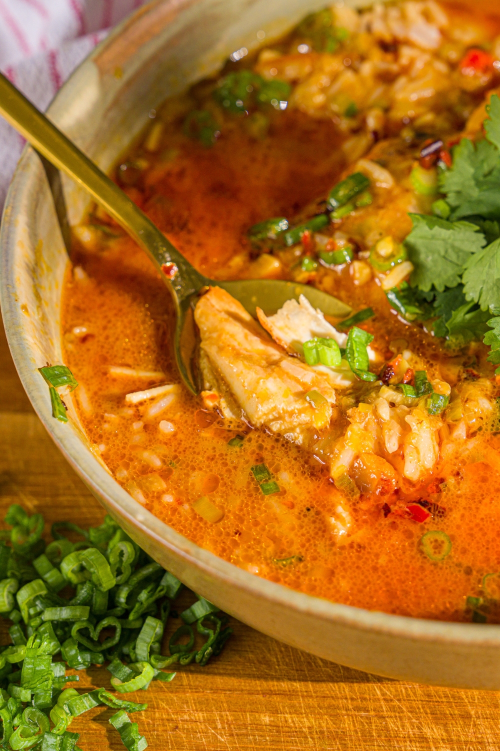 A bowl of thai coconut chicken bowl garnished with sliced green onion, cilantro, and chili oil. A spoon is dipped in the bowl. The bowl is on a wooden board.