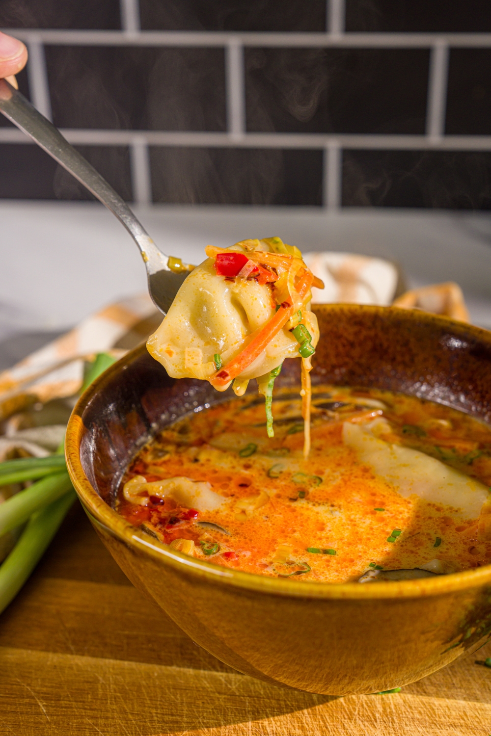 A bowl of creamy potsticker soup garnished with green onions and chili crisp. A spoon is taking a bite from the bowl. The bowl is on a wooden board with green onion.