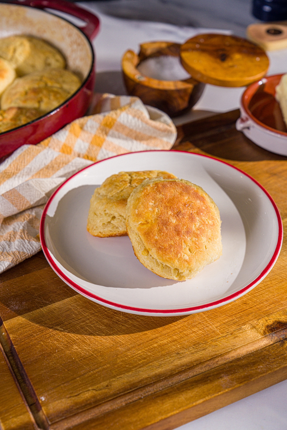 A plate with two Hardee's biscuits. The plate is on a wooden board with a yellow checkered napkin and baking dish of biscuits.