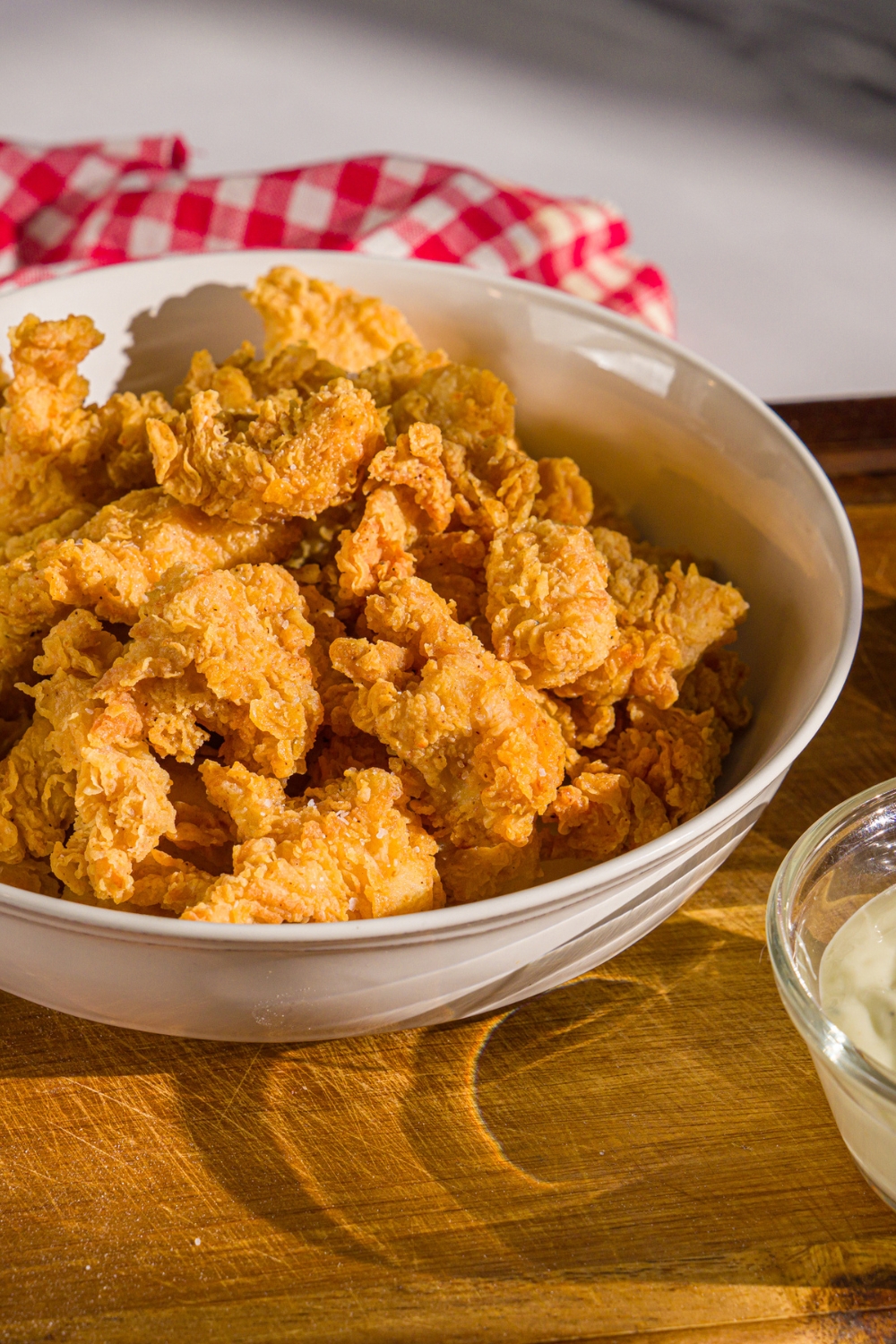A bowl of popcorn chicken on a wooden board with a bowl of dipping sauce. The board is on a marble counter with a red striped napkin.