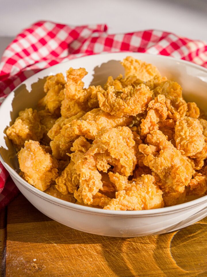 A bowl of popcorn chicken on a wooden board. The board is on a marble counter with a red striped napkin.