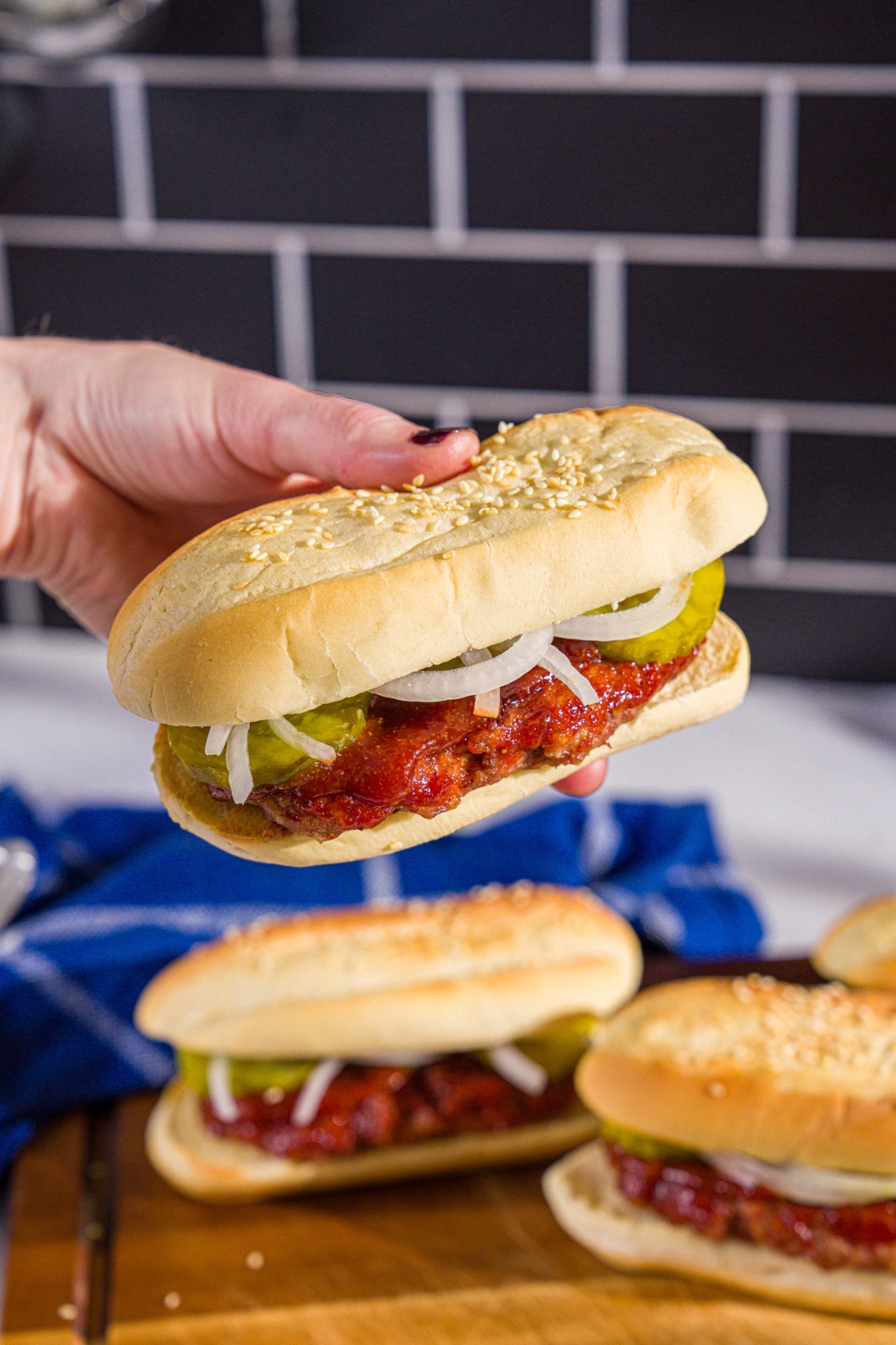 A close up of a hand holding a homemade McRib sandwich on a hoagie roll with pickles, onions, and homemade BBQ sauce. There is a wooden board with additional sandwiches blurred in the background.