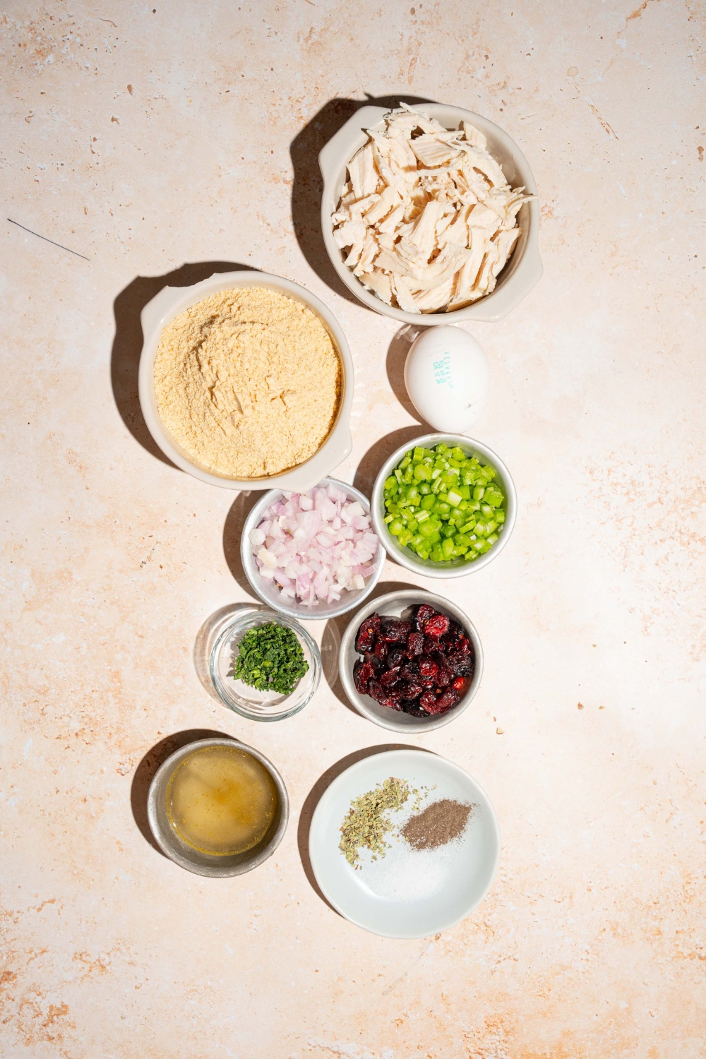 A tan counter with several bowls containing ingredients to make cranberry turkey stuffing balls including shredded turkey, bread crumbs, dried cranberries, onion, celery, egg, chicken broth, and seasonings.