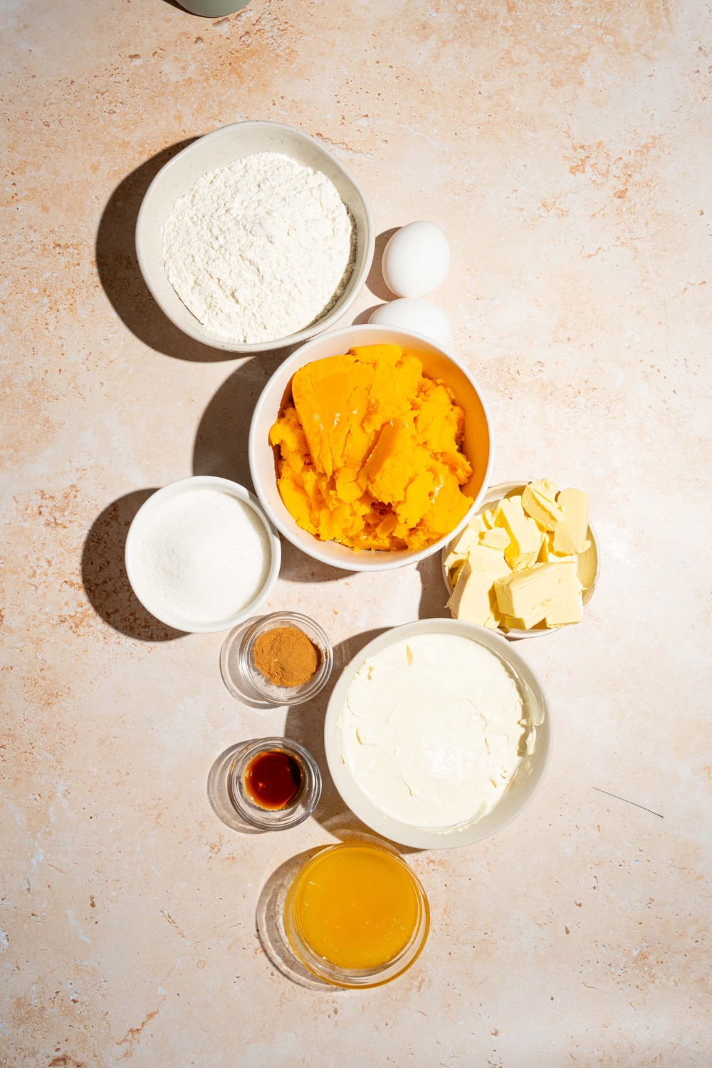 A tan counter with several bowls containing ingredients to make cream cheese pumpkin pie including cream cheese, pumpkin puree, sugar, vanilla, butter, egg, flour, and pumpkin pie spice.