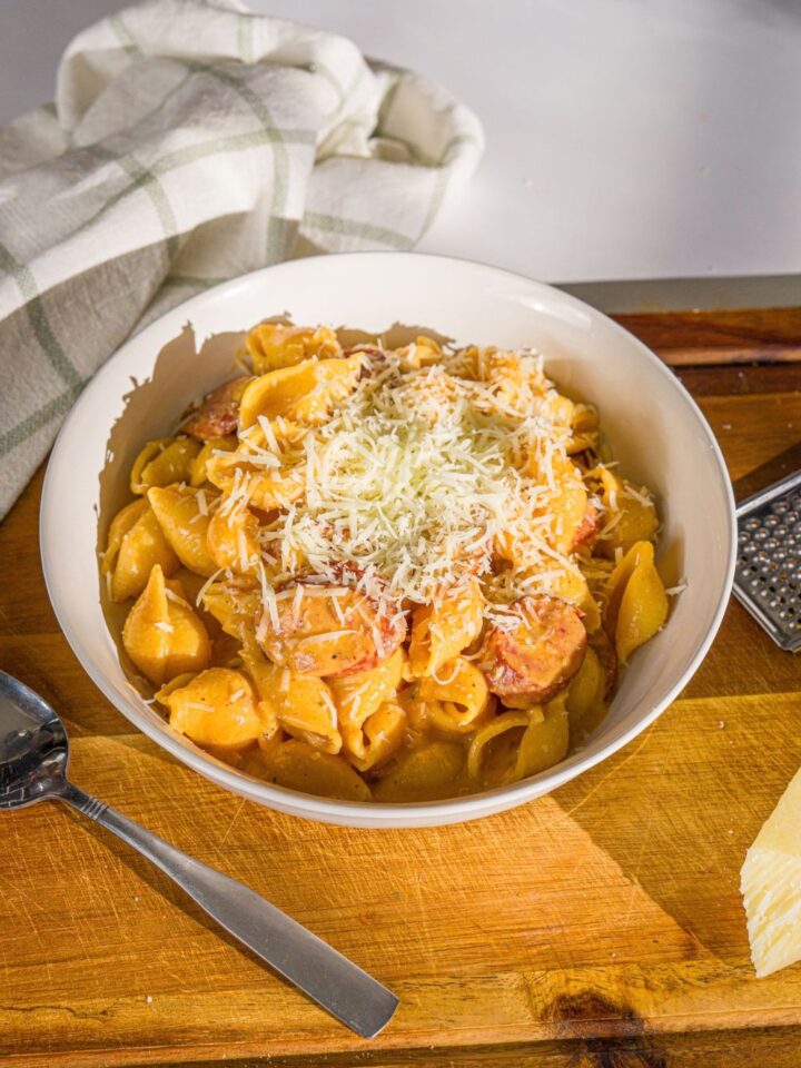 A white bowl with cajun sausage pasta topped with shredded parmesan cheese. The bowl is on a wooden board with a spoon and white cloth napkin.