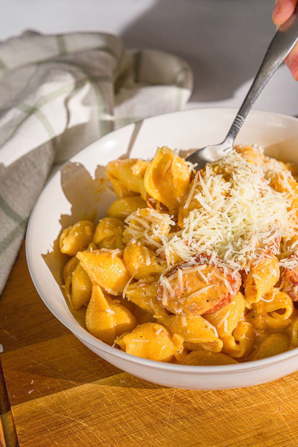 A white bowl with cajun sausage pasta topped with shredded parmesan cheese. A spoon is taking a bite from the bowl. The bowl is on a wooden board with a white cloth napkin.