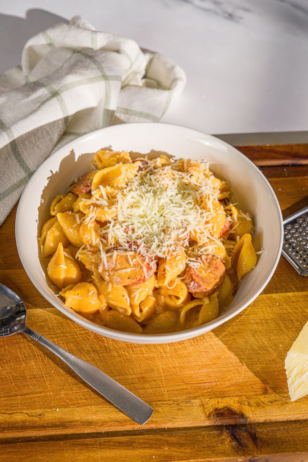 A white bowl with cajun sausage pasta topped with shredded parmesan cheese. The bowl is on a wooden board with a spoon and white cloth napkin.