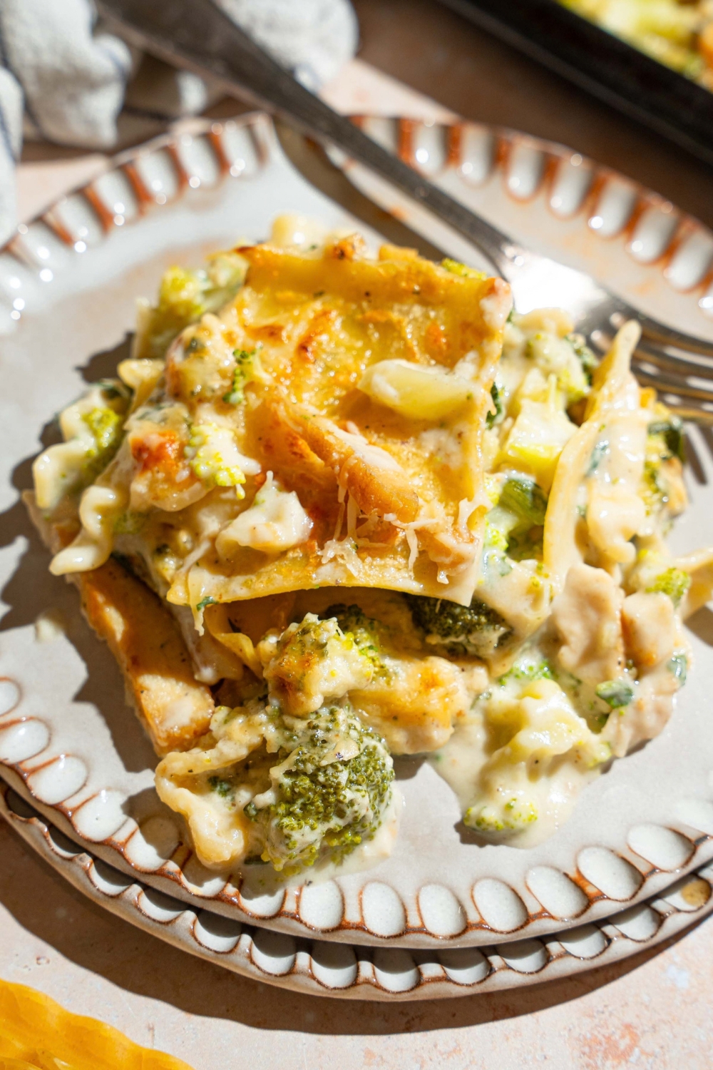 A plate with a slice of sheet pan chicken broccoli sheet pan lasagna. There is a fork on the plate. The plate is on a tan counter with a sheet pan of lasagna.