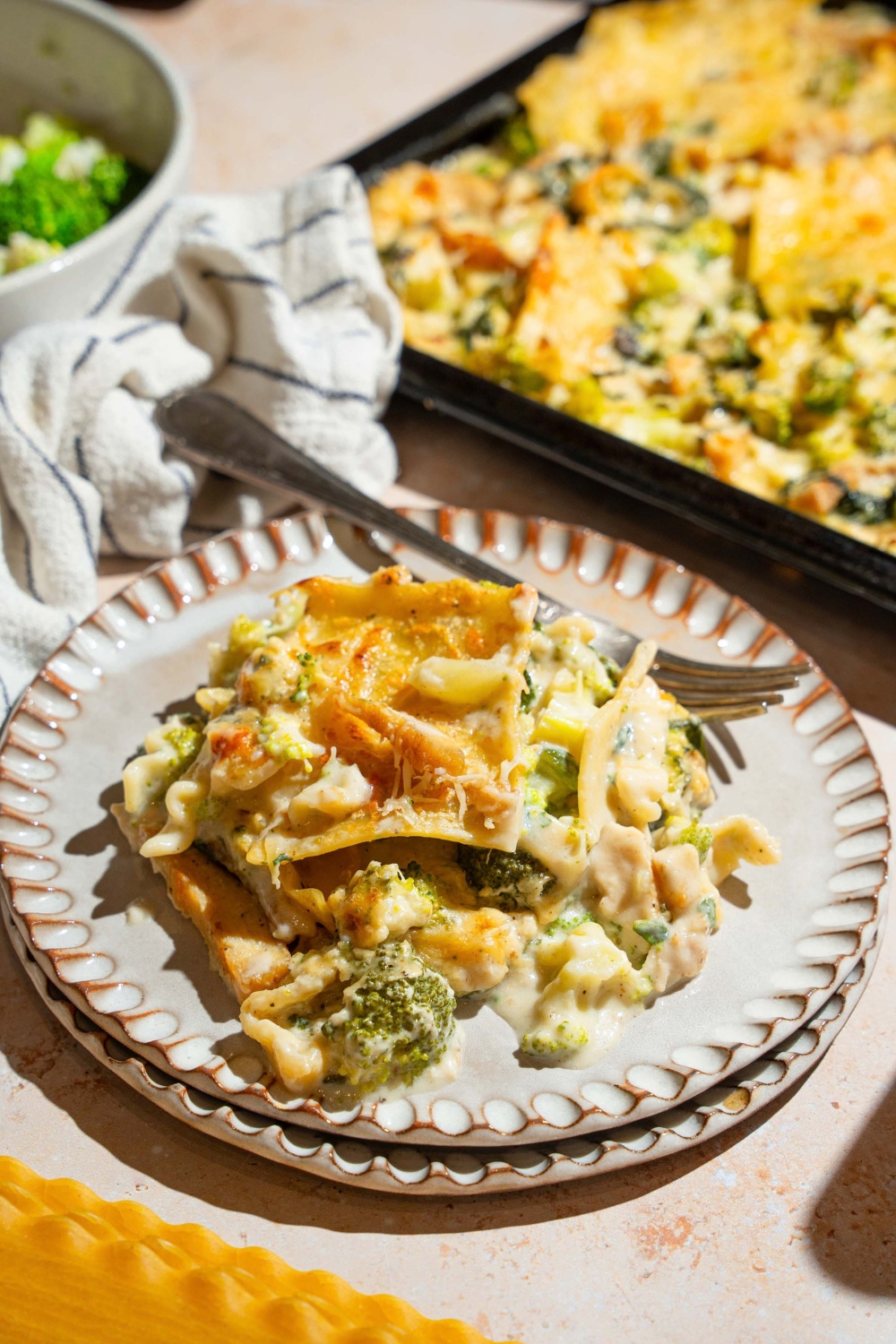 A plate with a slice of sheet pan chicken broccoli sheet pan lasagna. There is a fork on the plate. The plate is on a tan counter with a sheet pan of lasagna.