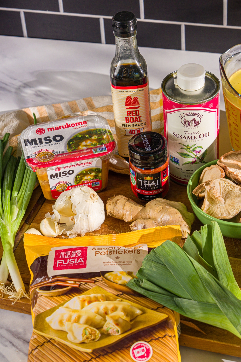 A wooden board with ingredients to make creamy potsticker soup including mushrooms, frozen potstickers, garlic, ginger, red curry, leeks, and more.