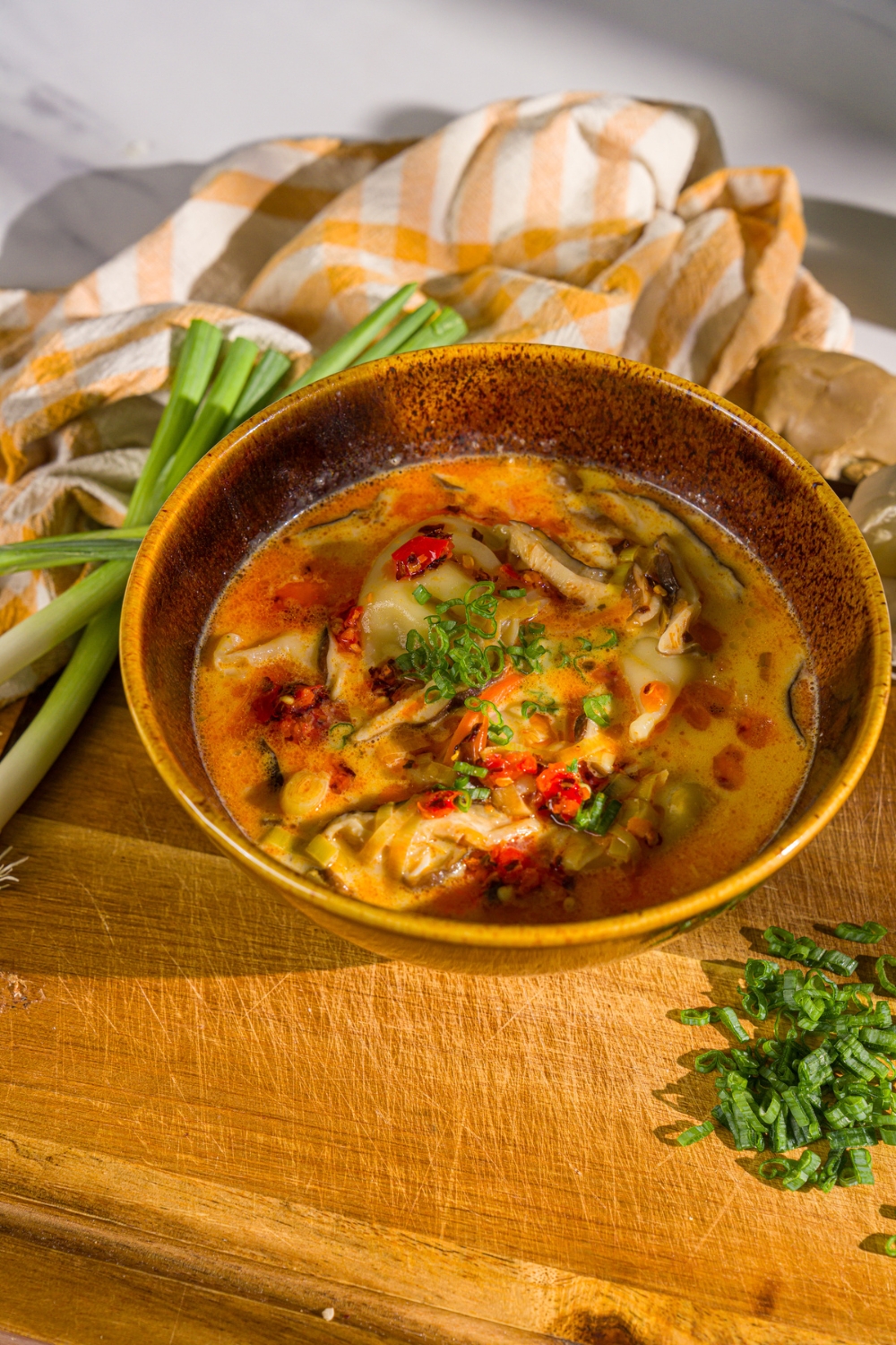A bowl of creamy potsticker soup garnished with green onions and chili crisp. The bowl is on a wooden board with green onion and a white cloth napkin.