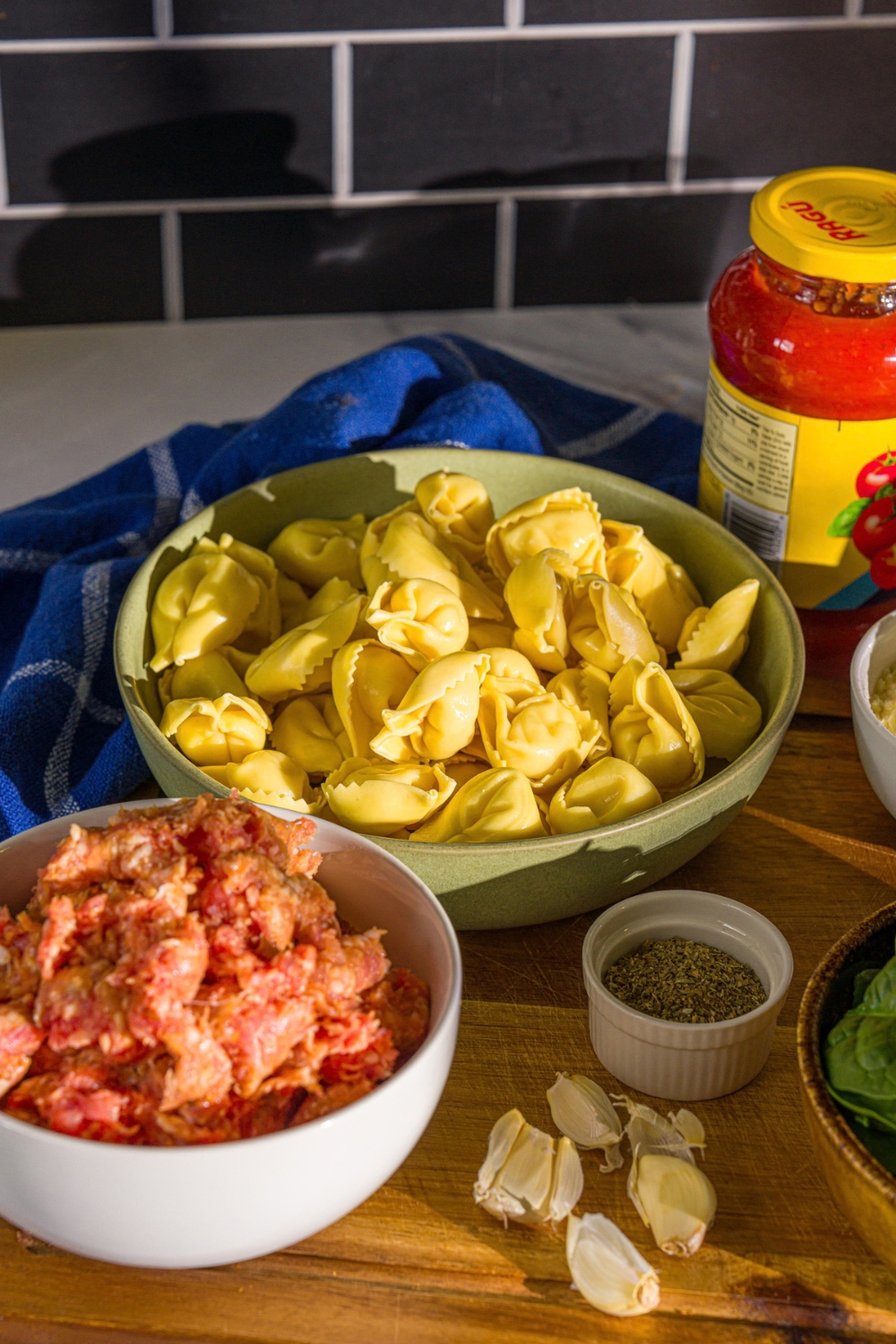 A wooden board with ingredients to make creamy tomato tortellini soup including ground sausage, tortellini, garlic, marinara sauce, and seasonings.