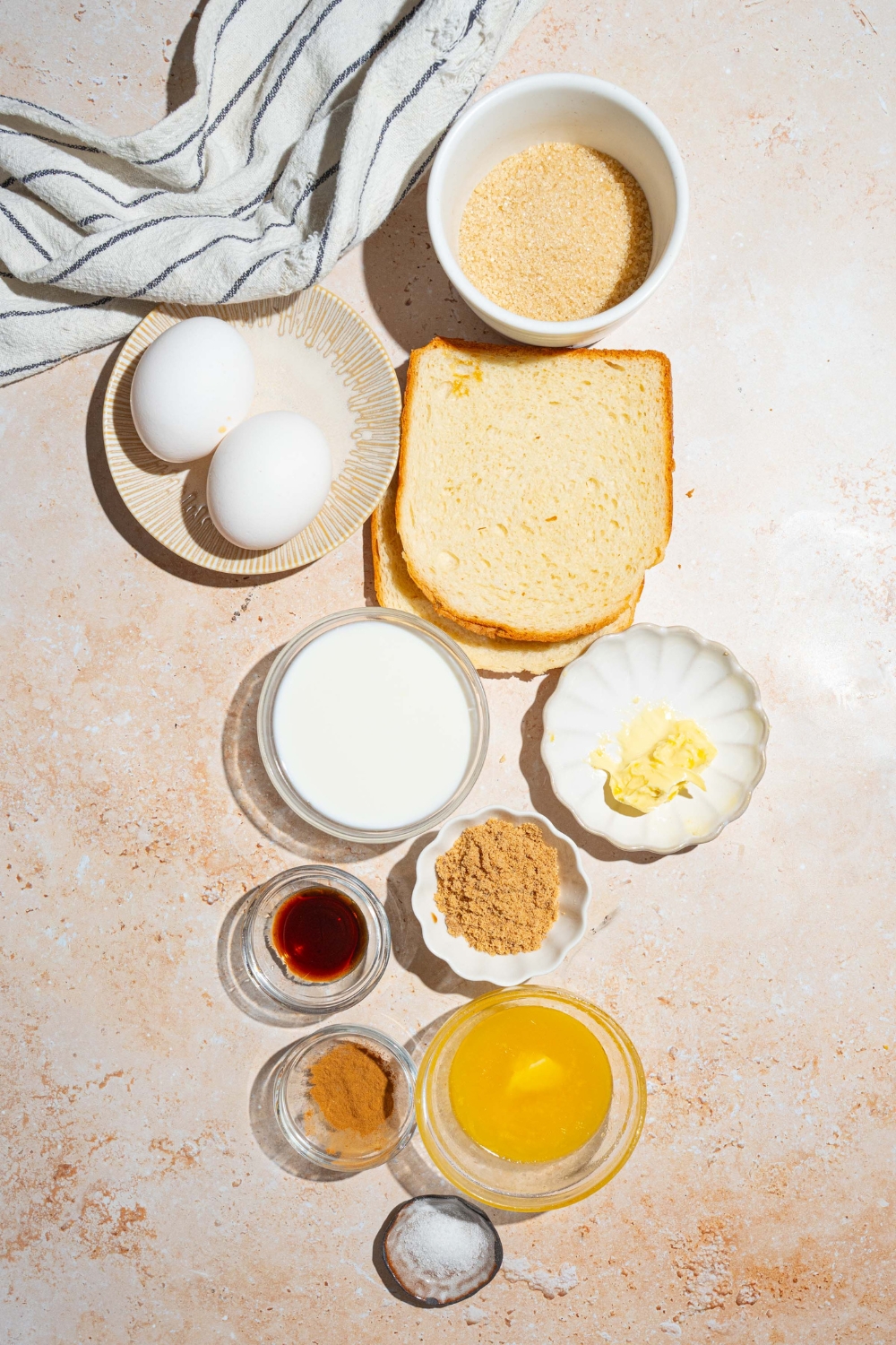 A tan counter with several bowls containing ingredients to make creme brulee french toast including eggs, sugar, butter, vanilla, milk, brioche bread, brown sugar, cinnamon, and more.